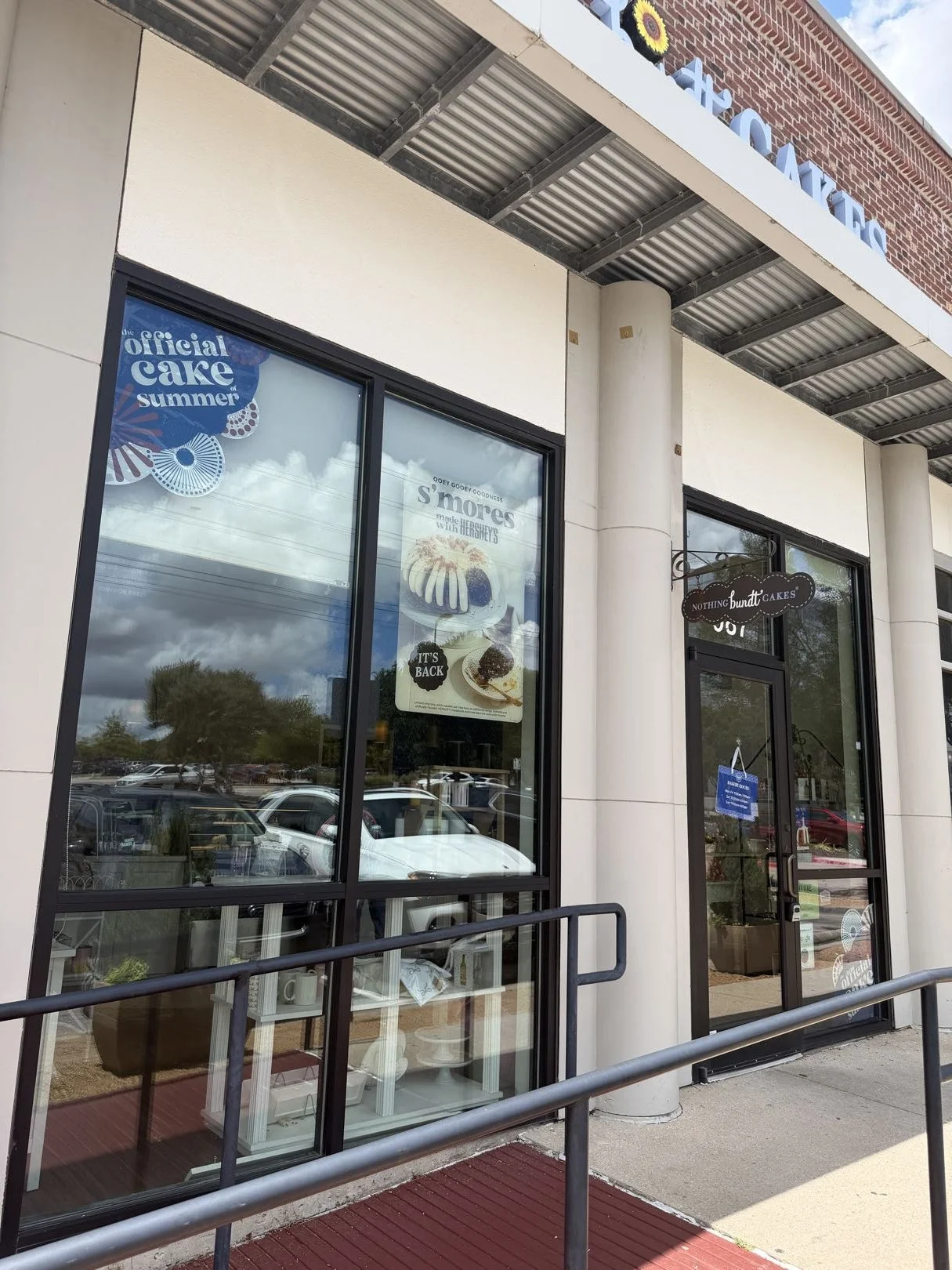 Exterior of a bakery shop with large glass windows and a door, promoting cakes and s'mores, with parking lot and cloudy sky reflected in the glass.
