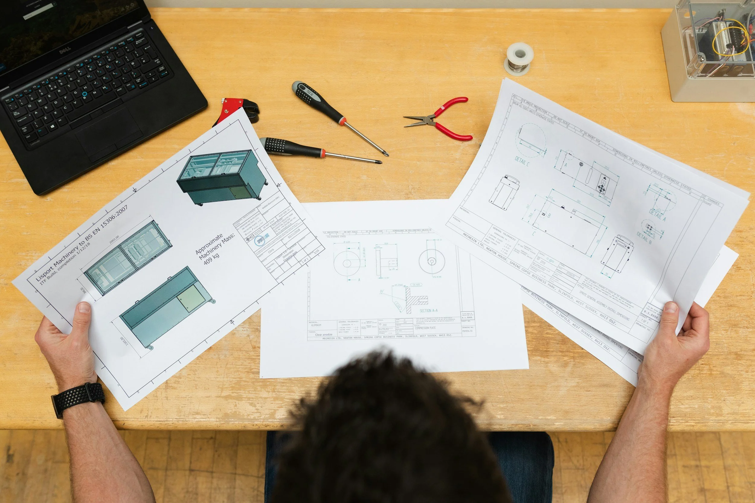 A person sitting at a wooden table reviewing technical drawings for a glass display case, with a Dell laptop, screwdrivers, a pair of pliers, a spool of wire, and an electrical component box on the table.