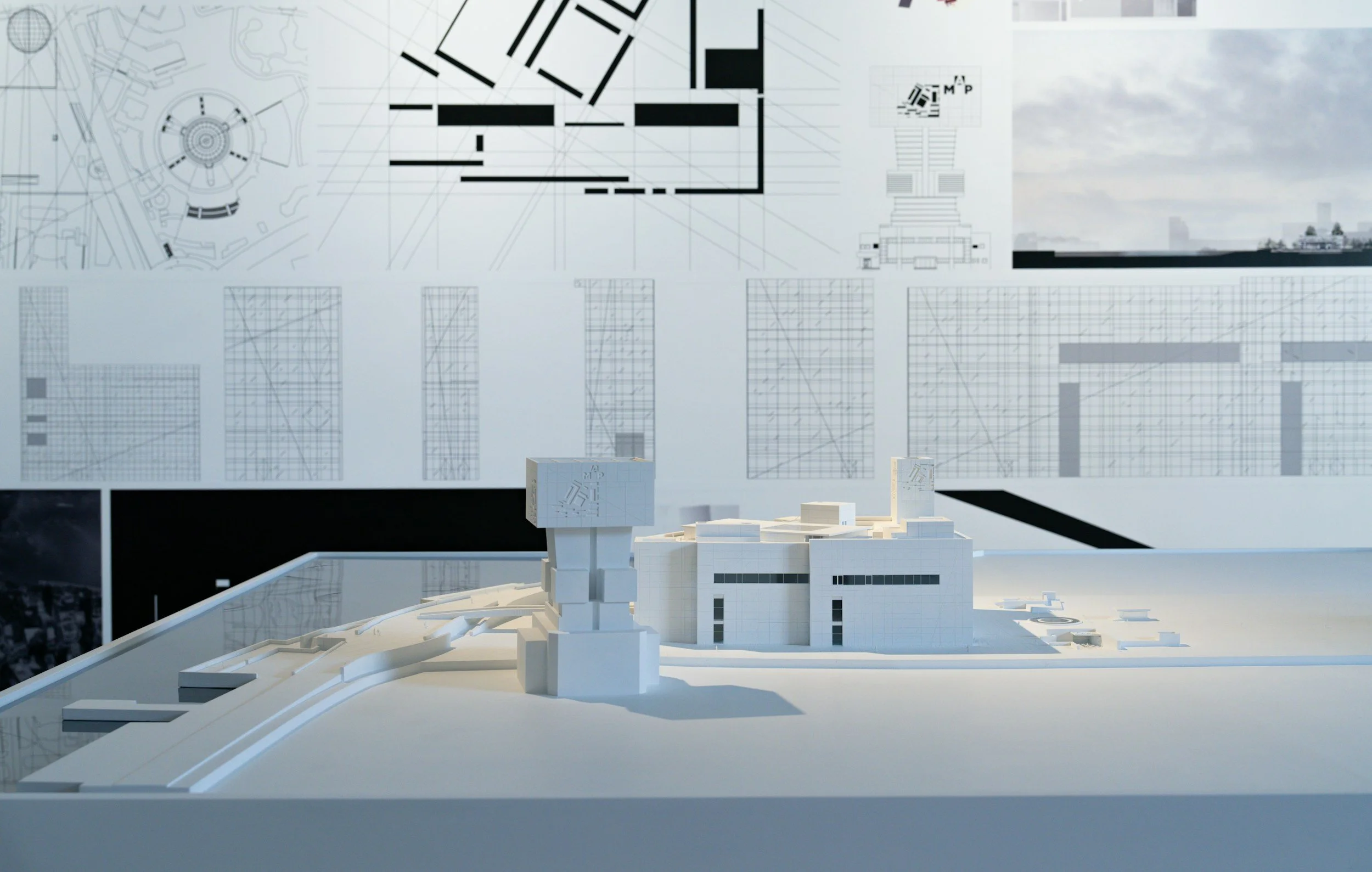 A detailed architectural model of a building complex with white structures, placed on a white surface. In the background, there is a large white wall featuring architectural drawings and plans, including floor layouts and elevations.