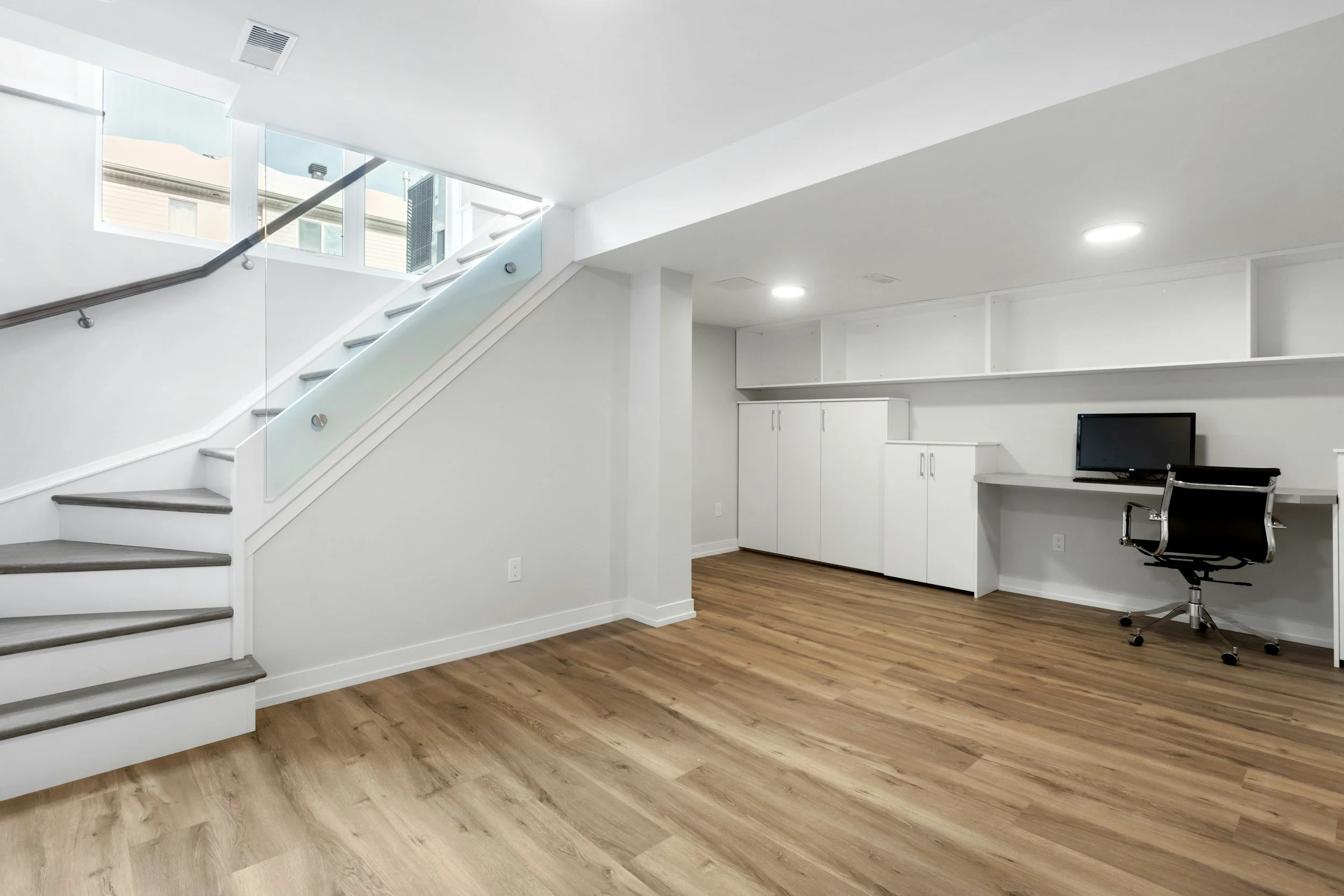 An empty modern office space with white walls, wooden flooring, a staircase with glass railing, a desk with a computer, and built-in cabinets.