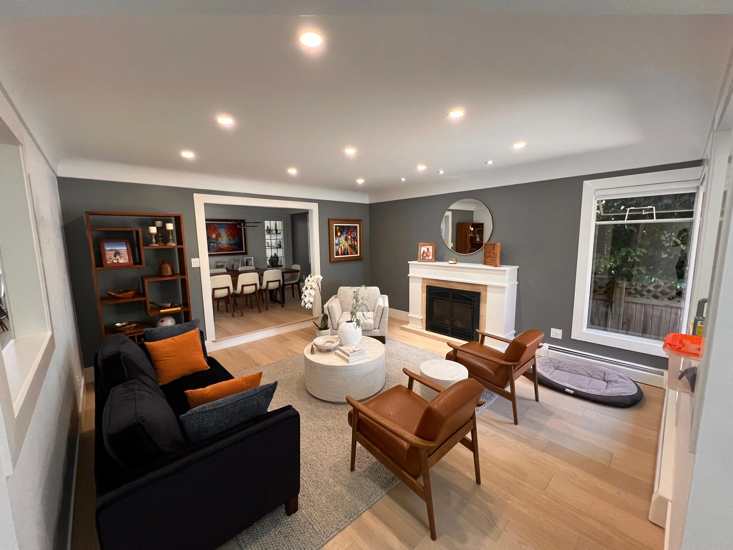 Living room with gray walls, hardwood floors, a black sofa with orange pillows, and wooden chairs. A fireplace with a round mirror above it, artwork on the walls, a dining area in the background, and a large window with a view outside.