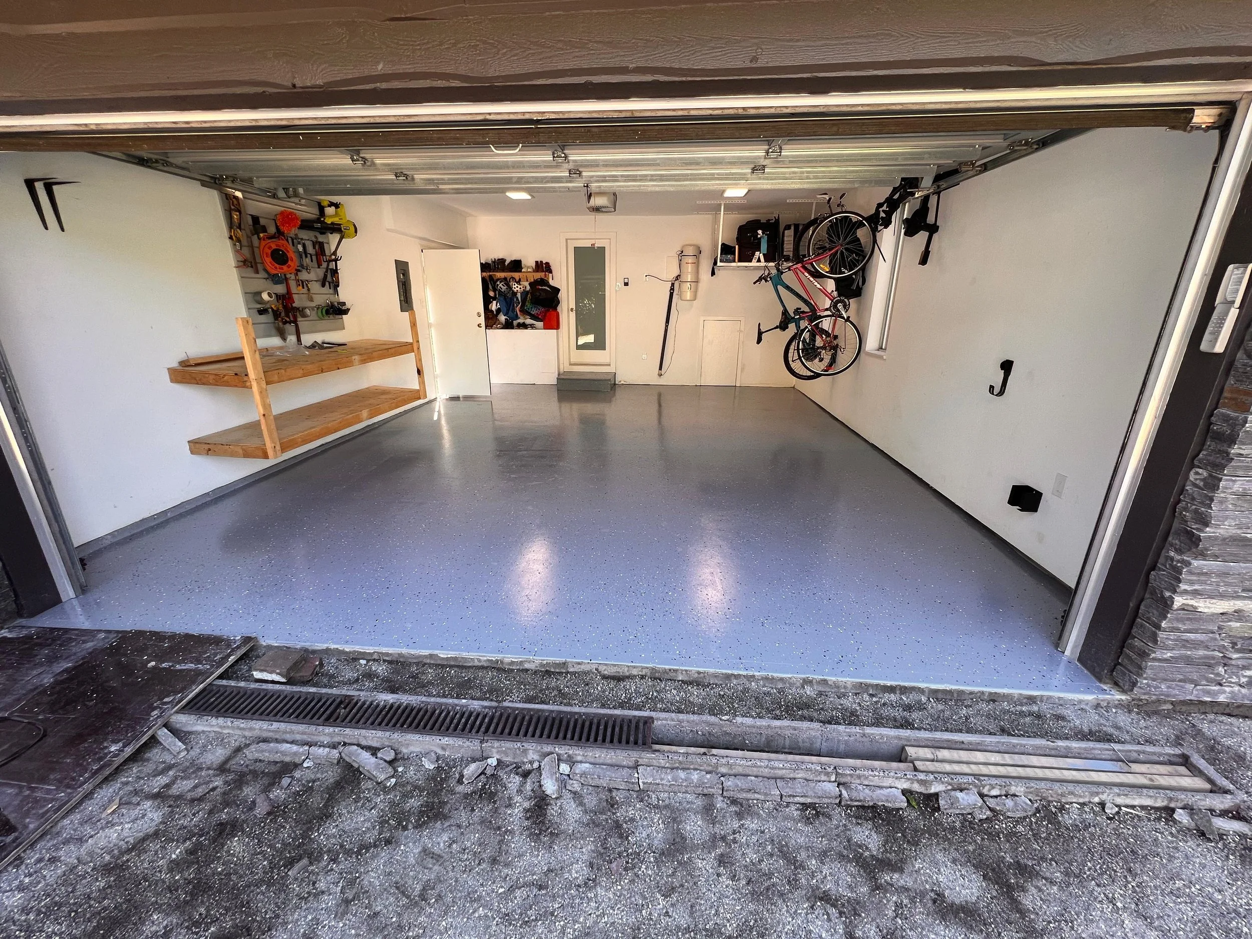 View of a garage with tools, bicycles, and a wooden workbench, with a partially open garage door showing the driveway outside.