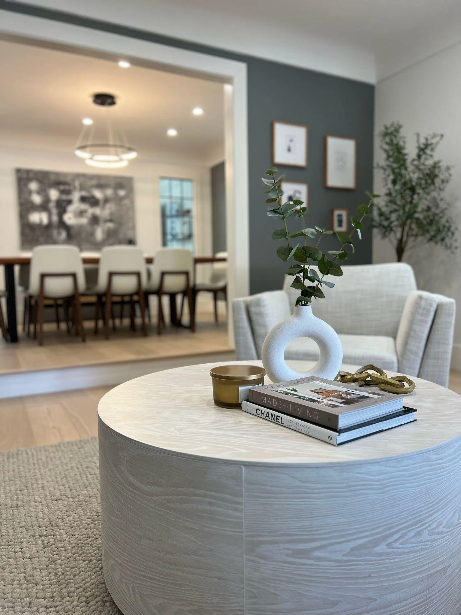 Close-up of a round, light-colored wooden coffee table with a white vase and green plant, a stack of books, and a gold cup in a modern living room.