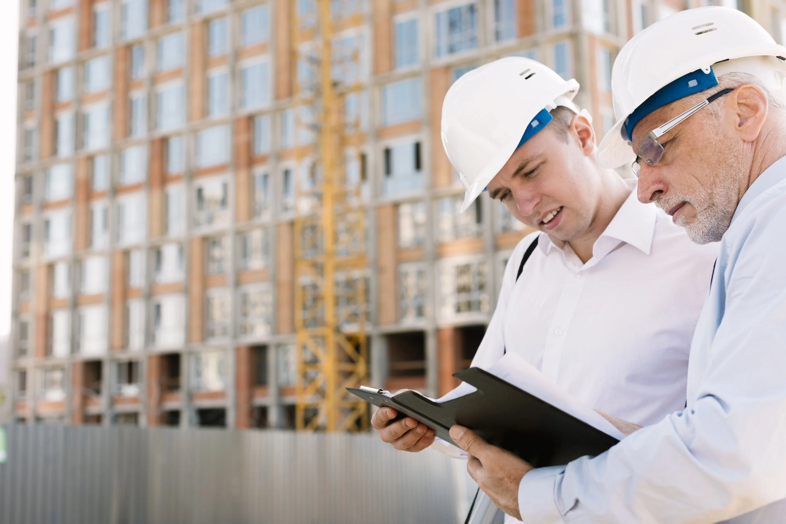 Two construction workers wearing white helmets and white shirts examining documents or plans at a construction site with building framework in the background.