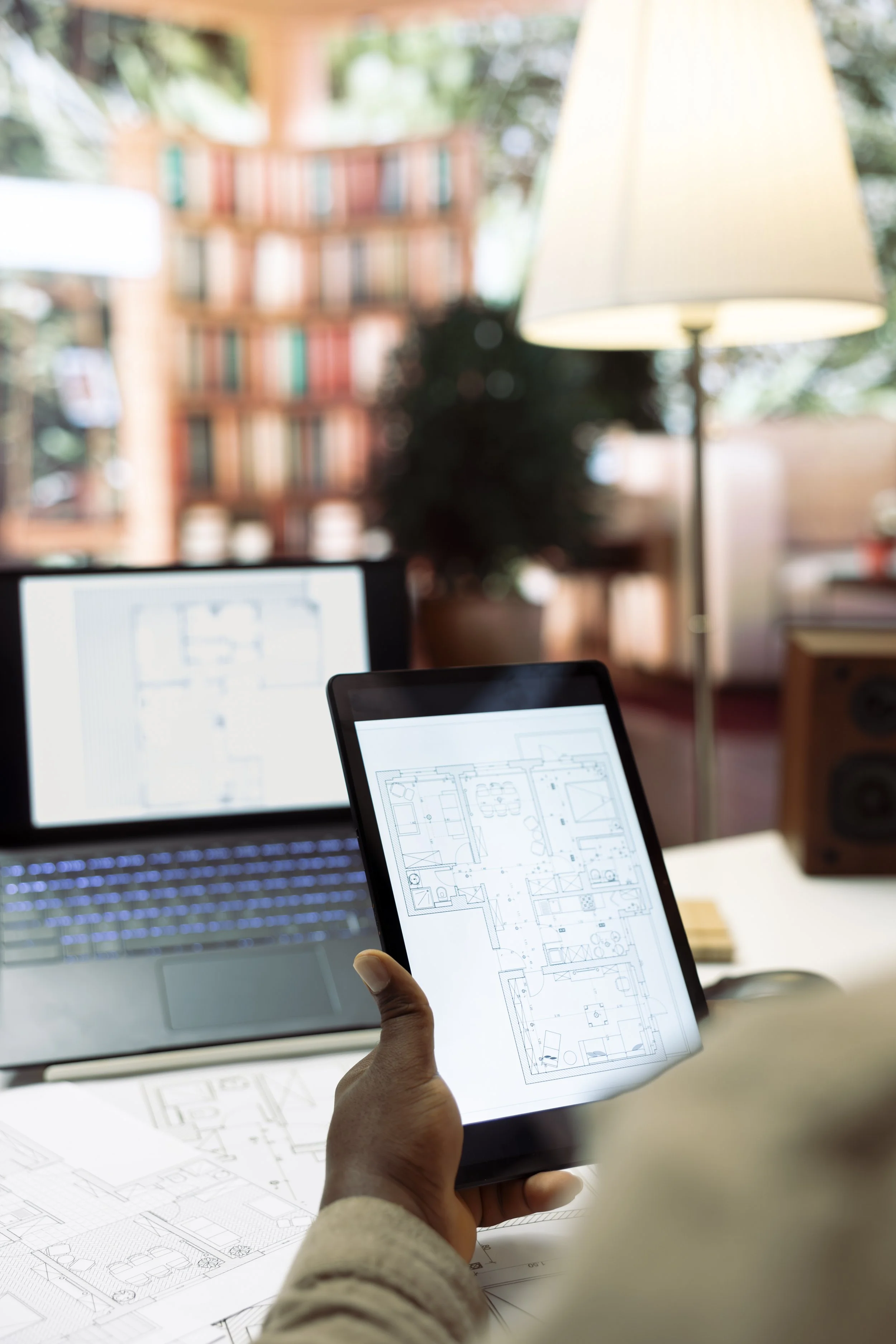 Person holding a tablet displaying a detailed home floor plan, with desks, monitors, and blueprints of architectural plans on a table in a well-lit room.