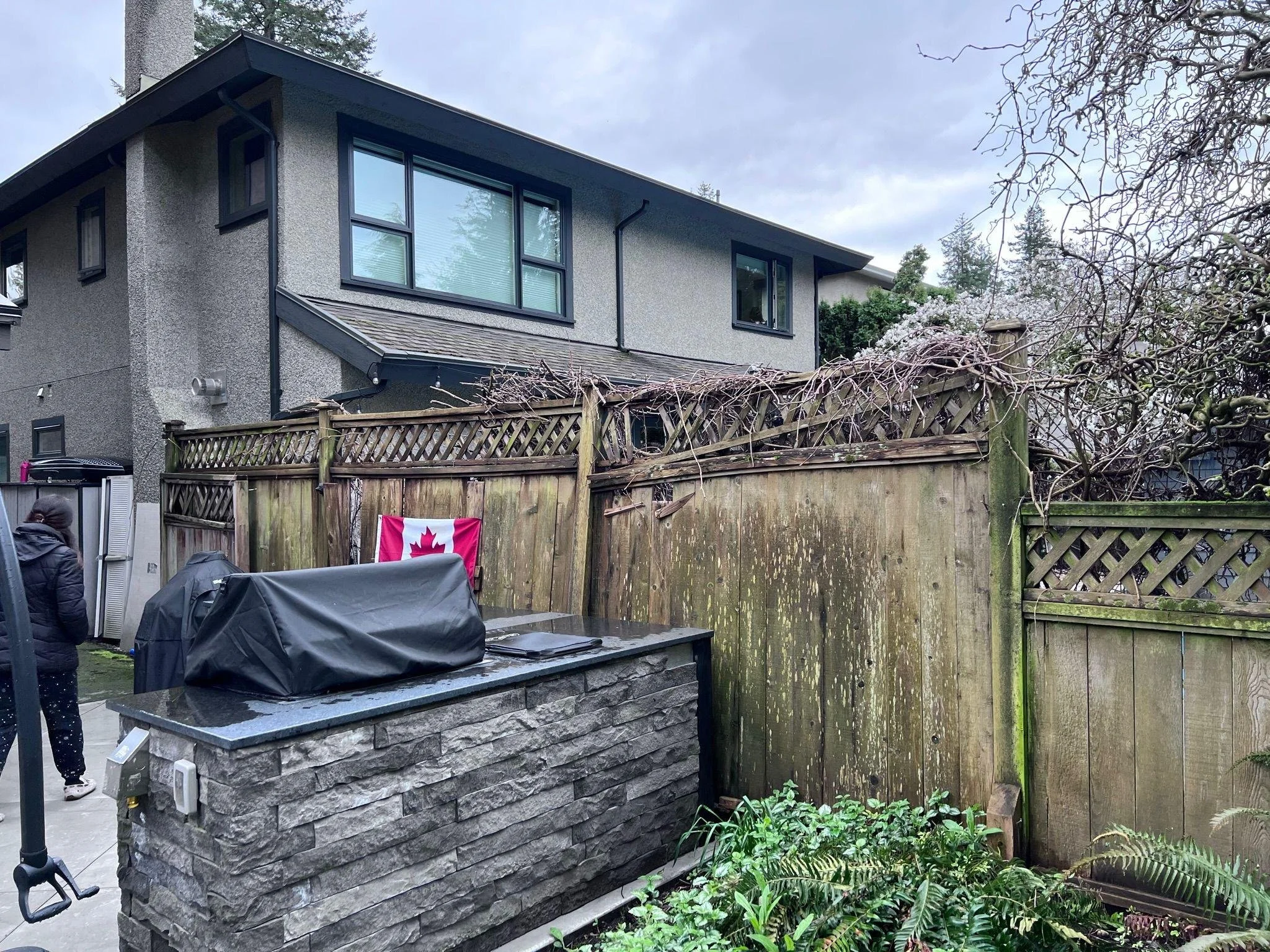 Rear view of a two-story house with large windows, surrounded by trees and a wooden fence. There is a stone outdoor fireplace or grill with a black cover in the foreground, and a Canadian flag hanging on the fence.