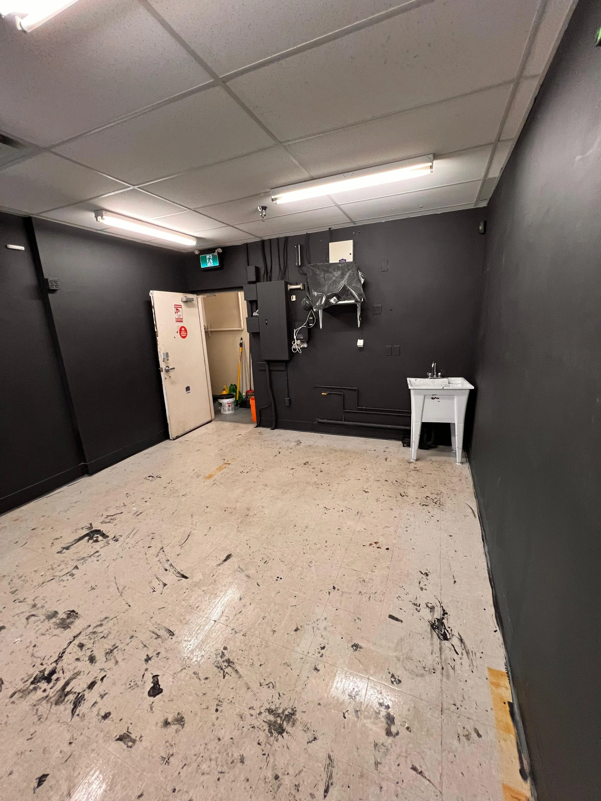 A room with a dark wall, fluorescent lighting, a small white sink, and a door partially open. The floor is covered with black smudges and debris.
