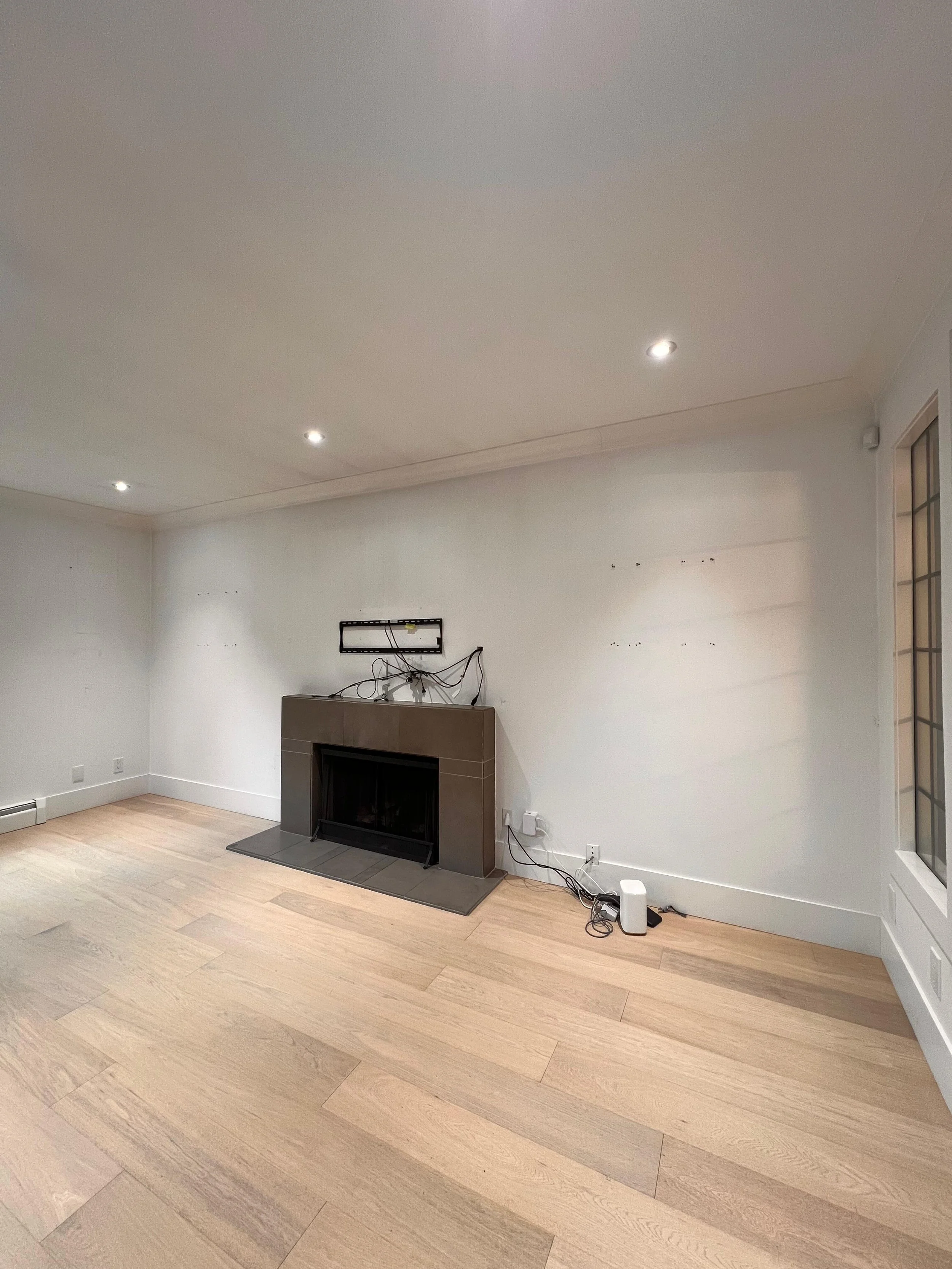 A living room with a beige brick fireplace and a mounted TV bracket on a white wall. The room has light-colored wooden flooring and a corner window with a grid pattern. Various electrical wires and devices are on the floor near the wall.