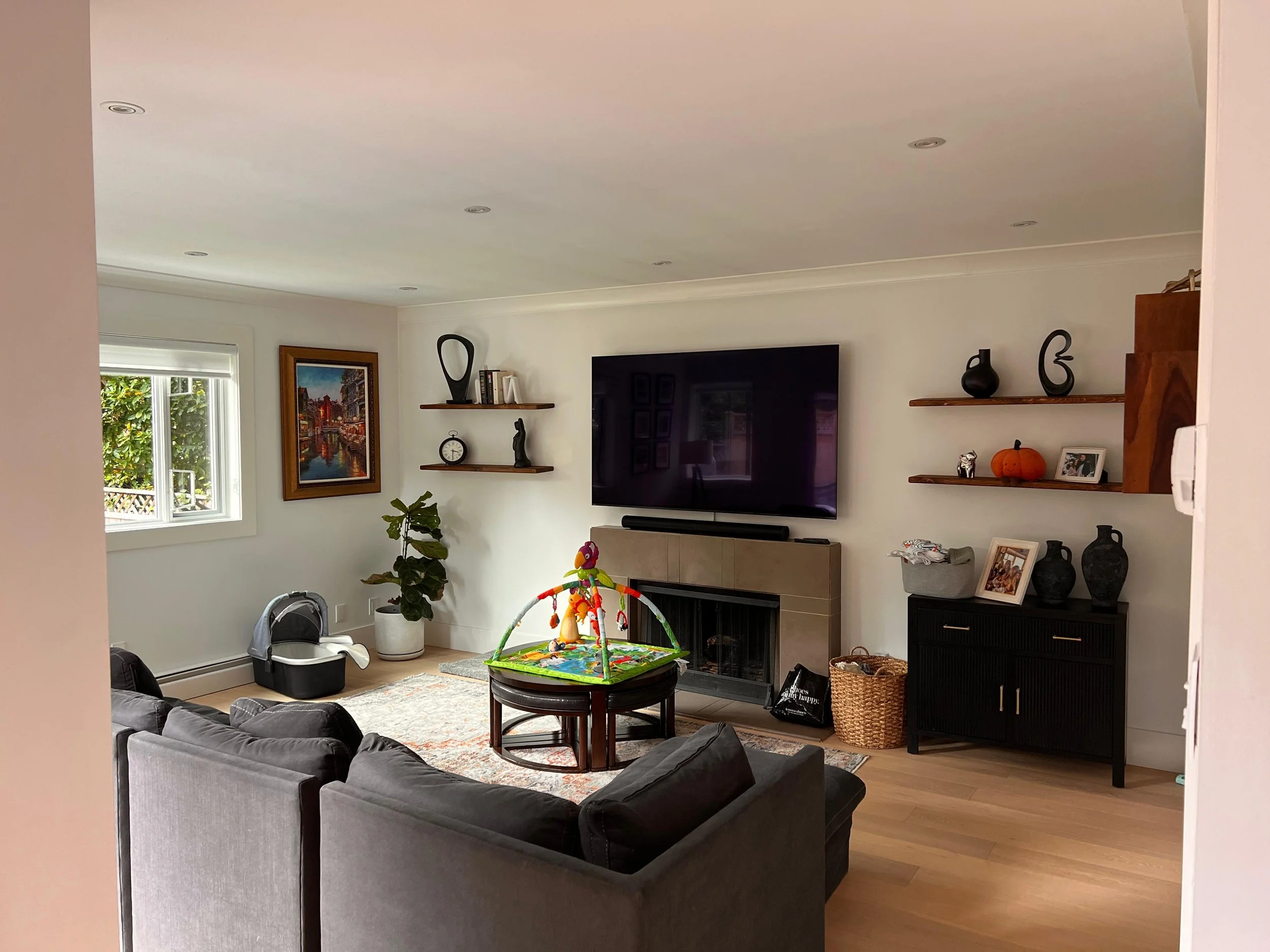Living room with grey sofa, round wooden table with colorful baby play mat, TV mounted on wall, two wooden shelves with decorative items, window with white blinds, framed painting of city scene, potted plant, and other household items.