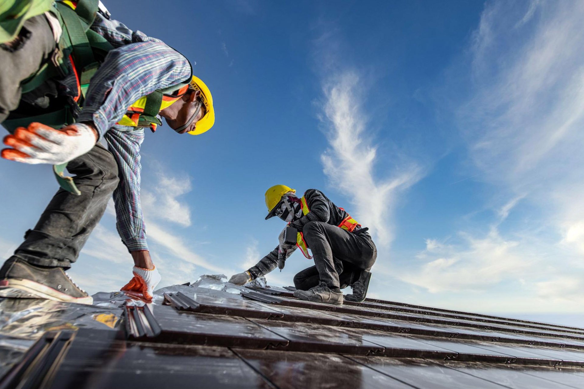 Three construction workers wearing safety helmets and vests are laying metal roofing sheets on a roof under a blue sky with some clouds.