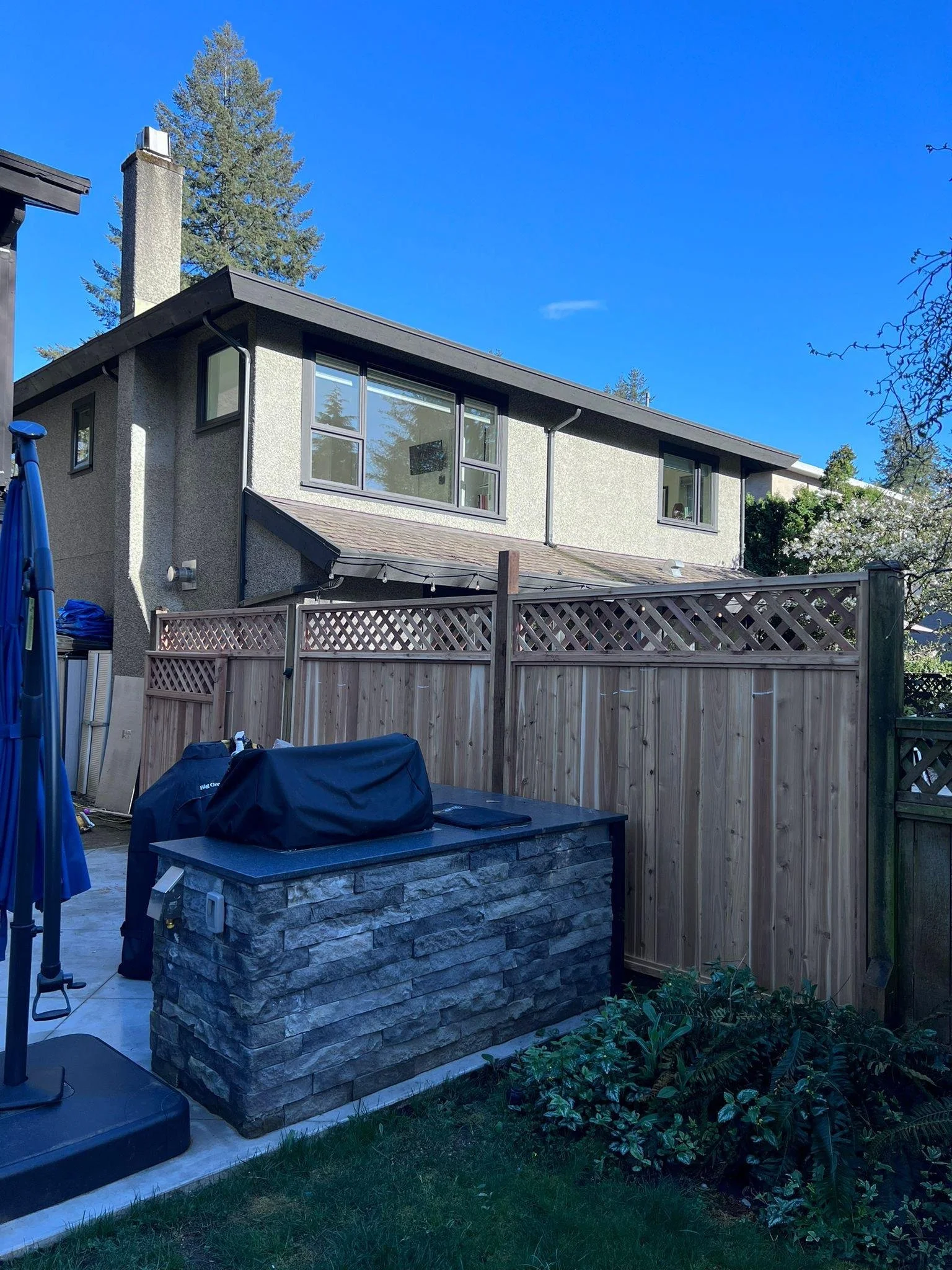 Backyard with a stone firepit, a wooden privacy fence, and a two-story house with large windows and a chimney, under a clear blue sky with tall trees in the background.