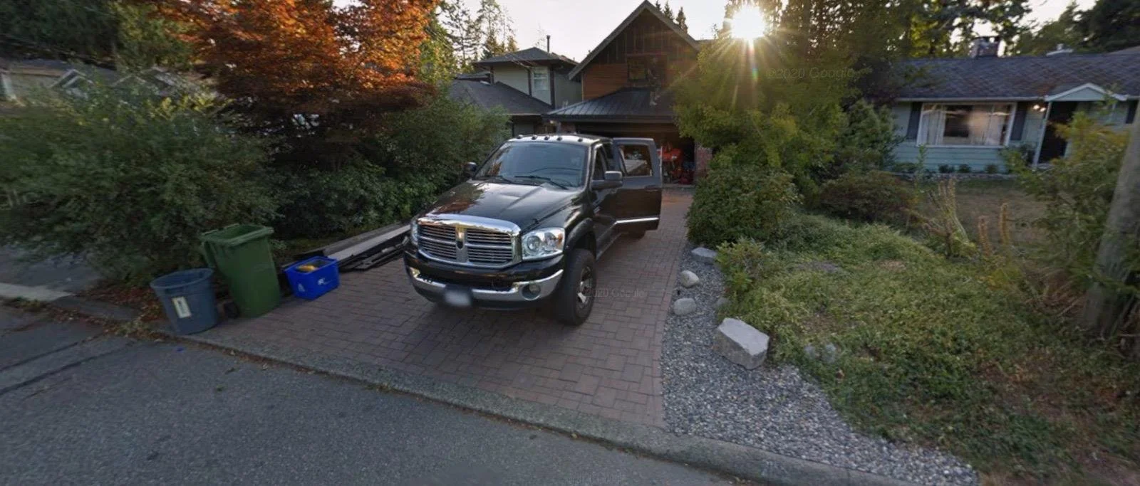 A black pickup truck is parked on a brick driveway in front of a house, with its front to the viewer. The driveway is bordered by a garden with bushes and trees. There are several garbage and recycling bins on the side of the driveway. The sun is setting behind the house, casting a glow over the scene.