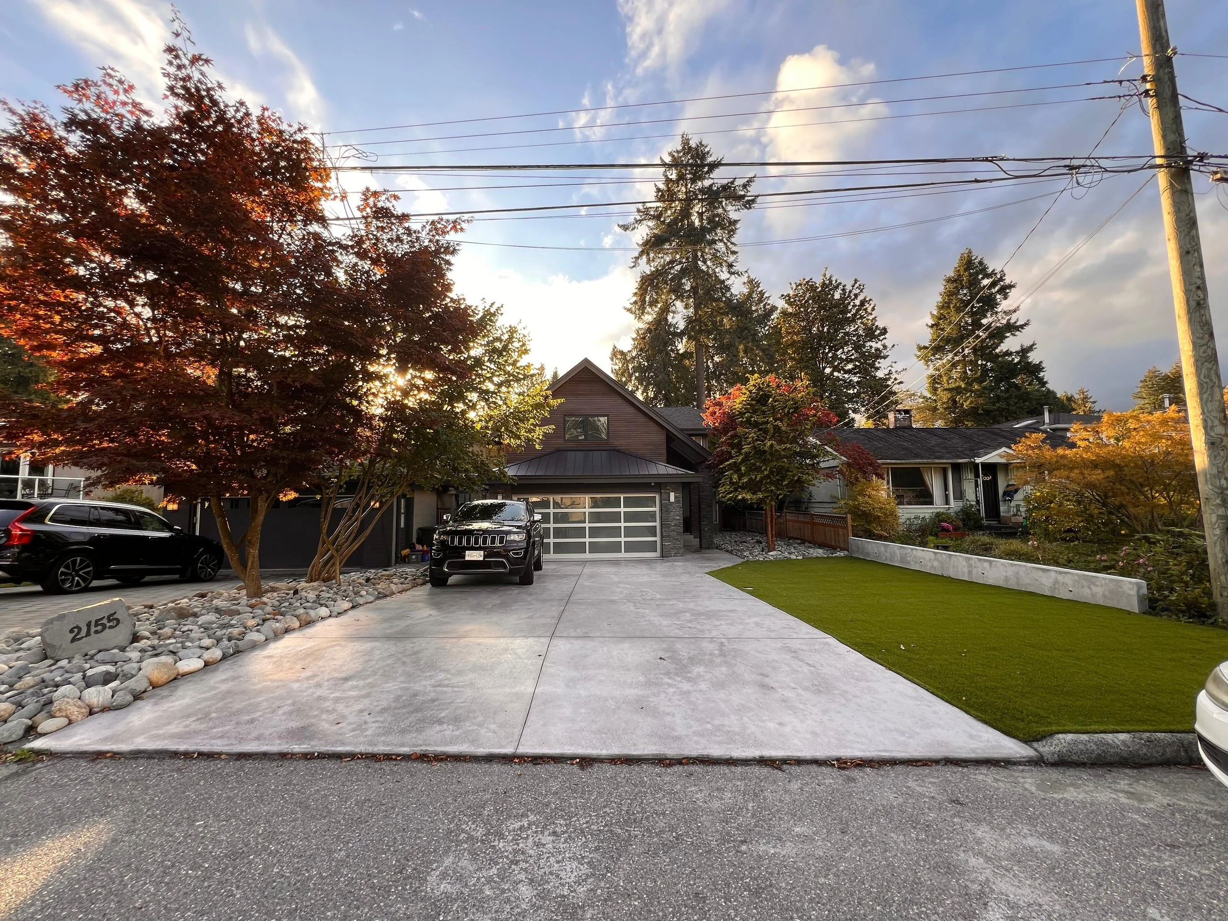 Residential driveway with a black SUV parked in front of a modern house surrounded by trees with autumn-colored leaves; blue sky with clouds and power lines overhead.