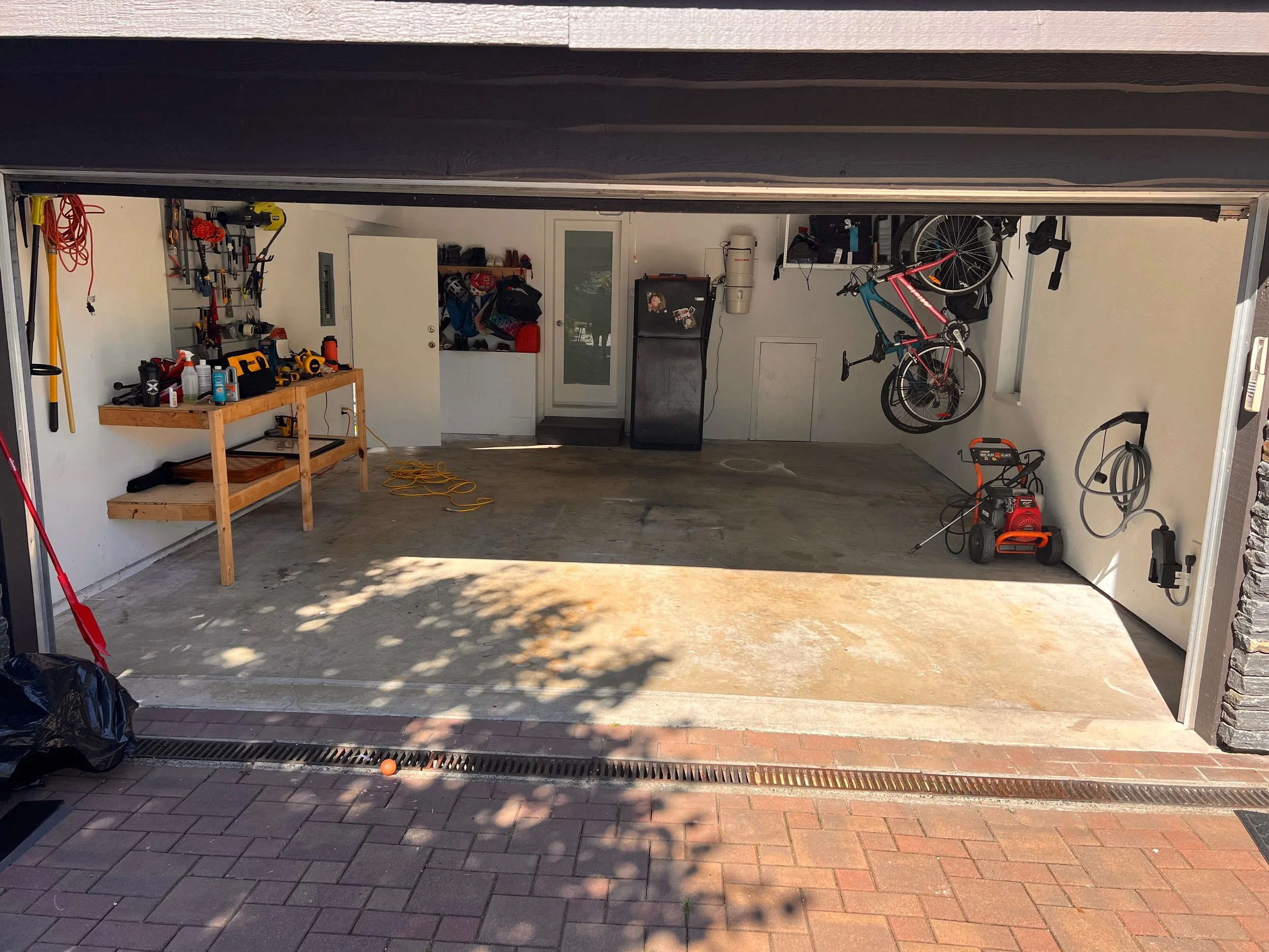The photo shows the inside of a garage with a bicycle hanging on the wall, a workbench with tools, and various items stored on shelves and in bins. The garage door is open, revealing a paved driveway outside.