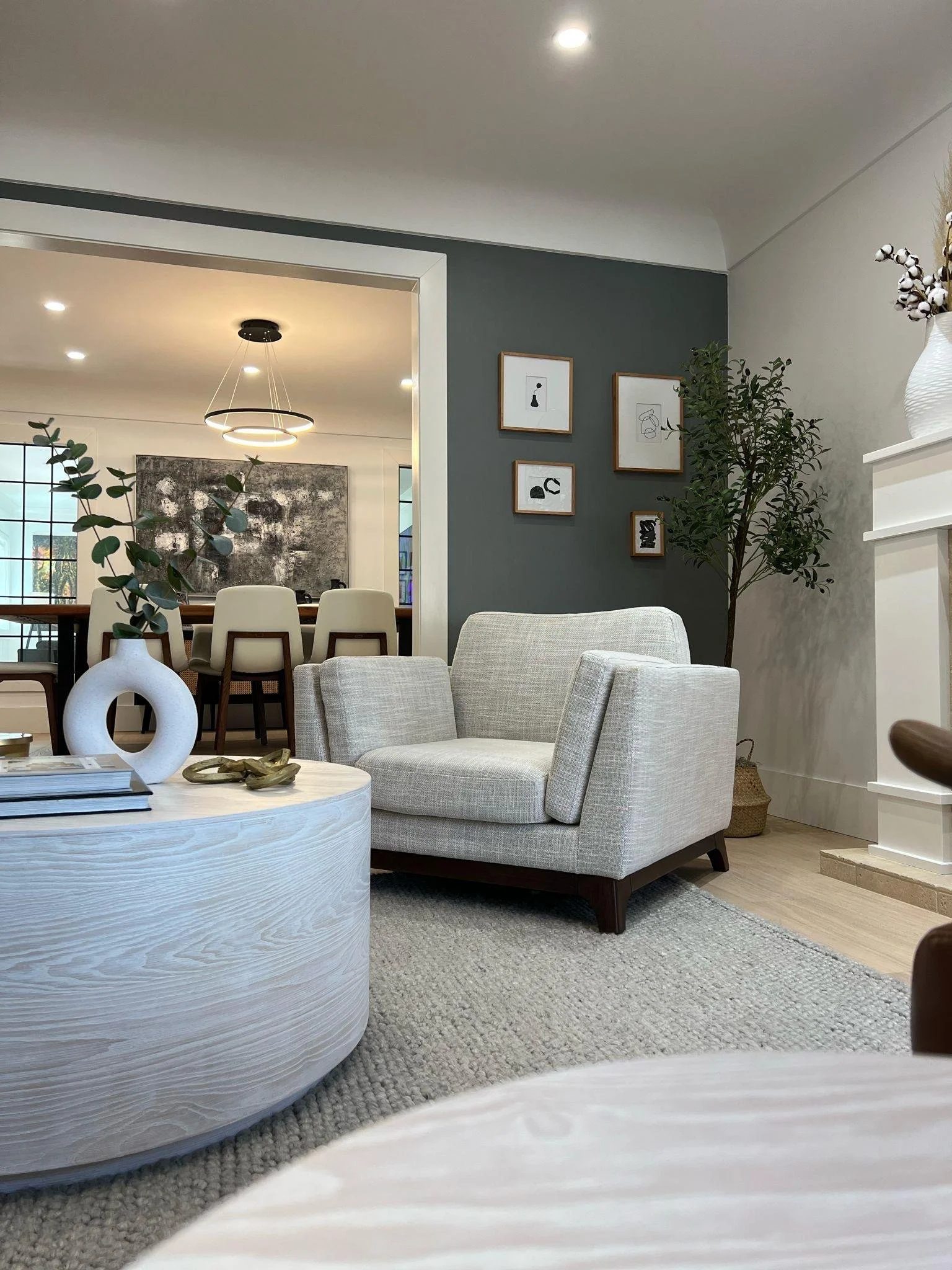 A modern living room with beige and gray furniture, wall art, a large window with multiple panes, decorative plants, and a textured light-colored rug.