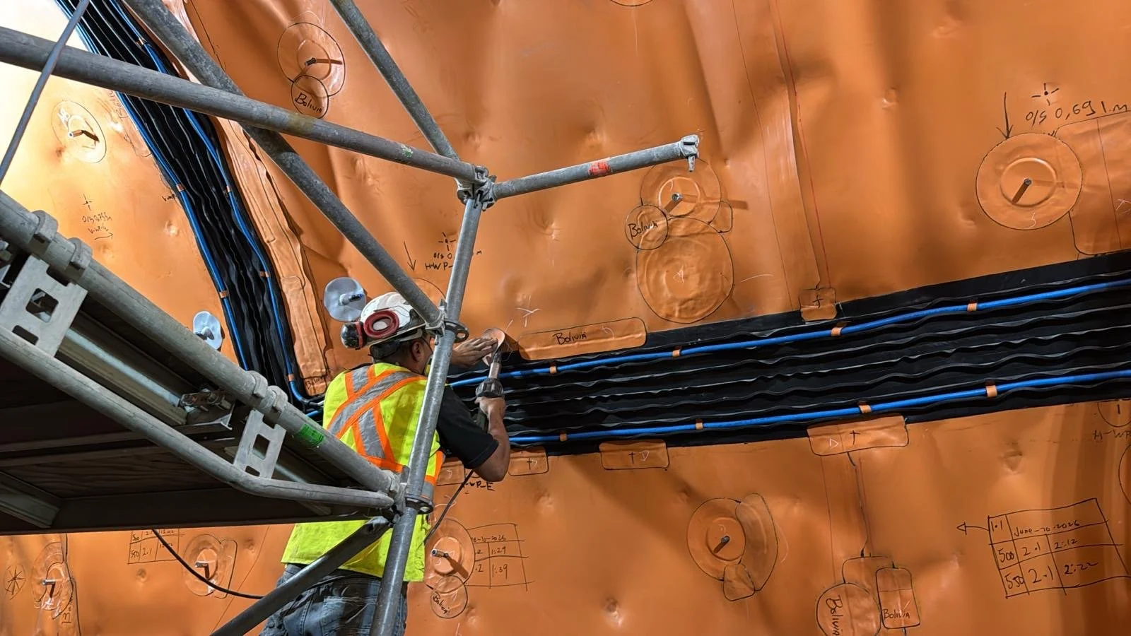 Construction workers on a building site, wearing orange safety vests and helmets, working among metal rebar and construction materials.
