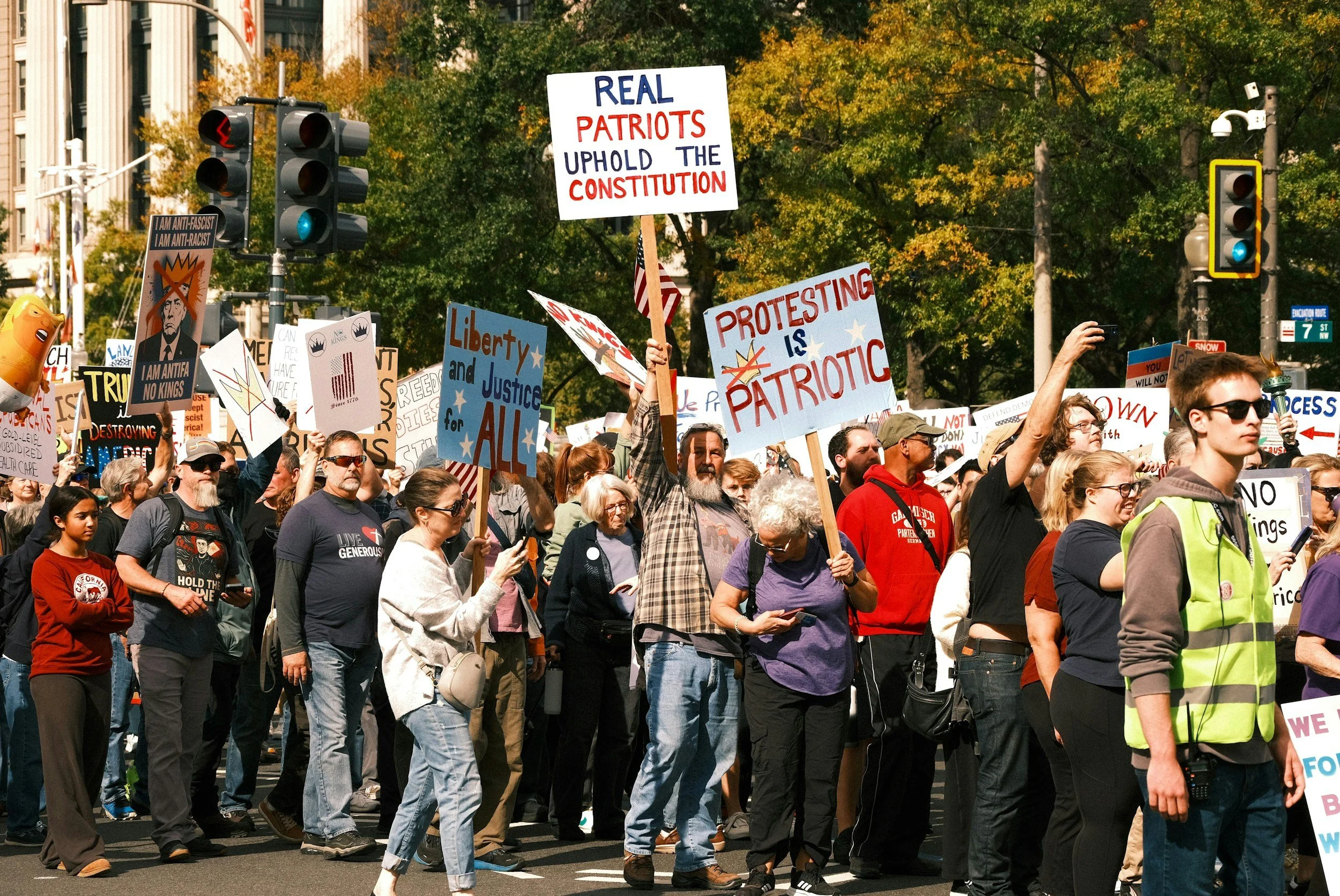 Crowd of protesters holding signs during a march or demonstration on a city street, with traffic lights and trees in the background.
