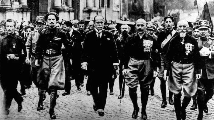 Group of men marching on cobblestone street, some wearing military uniforms with medals, others in suits, in black and white photograph.