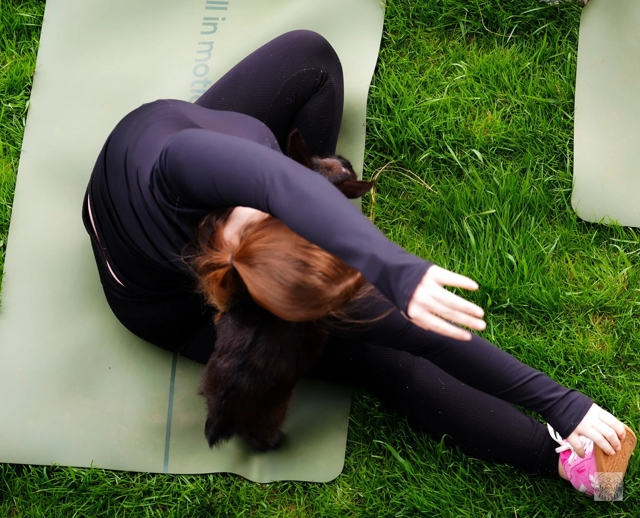 A person is lying on a green exercise mat outdoors on grass, holding a small dog against their chest, with another person's foot visible in a pink sneaker nearby.
