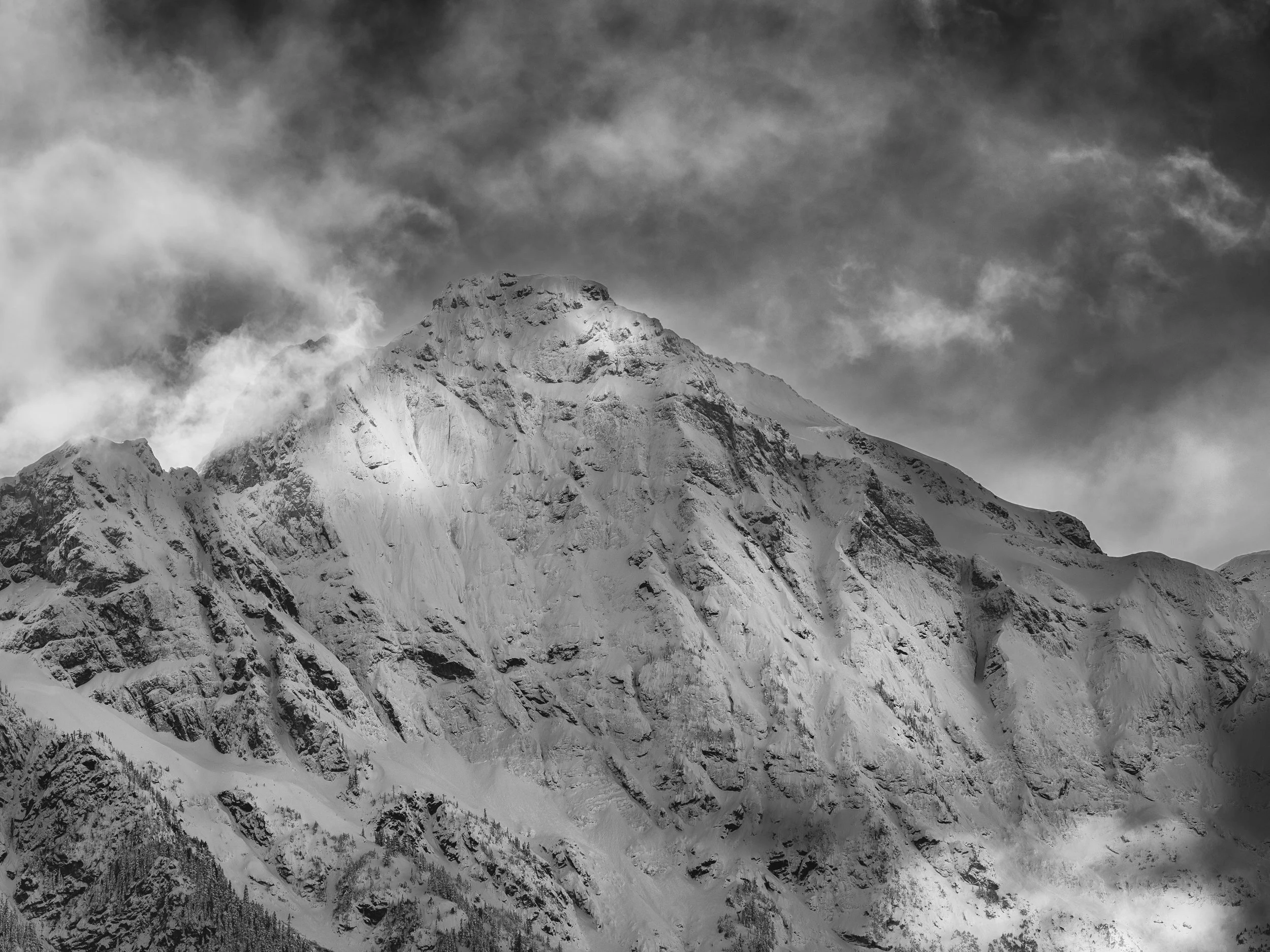 A shaft of storm light has found the mountain's central face and illuminated it while leaving everything around it in darker register — the flanking ridges, the lower slopes, the turbulent clouds above. The effect is something between spotlight and r