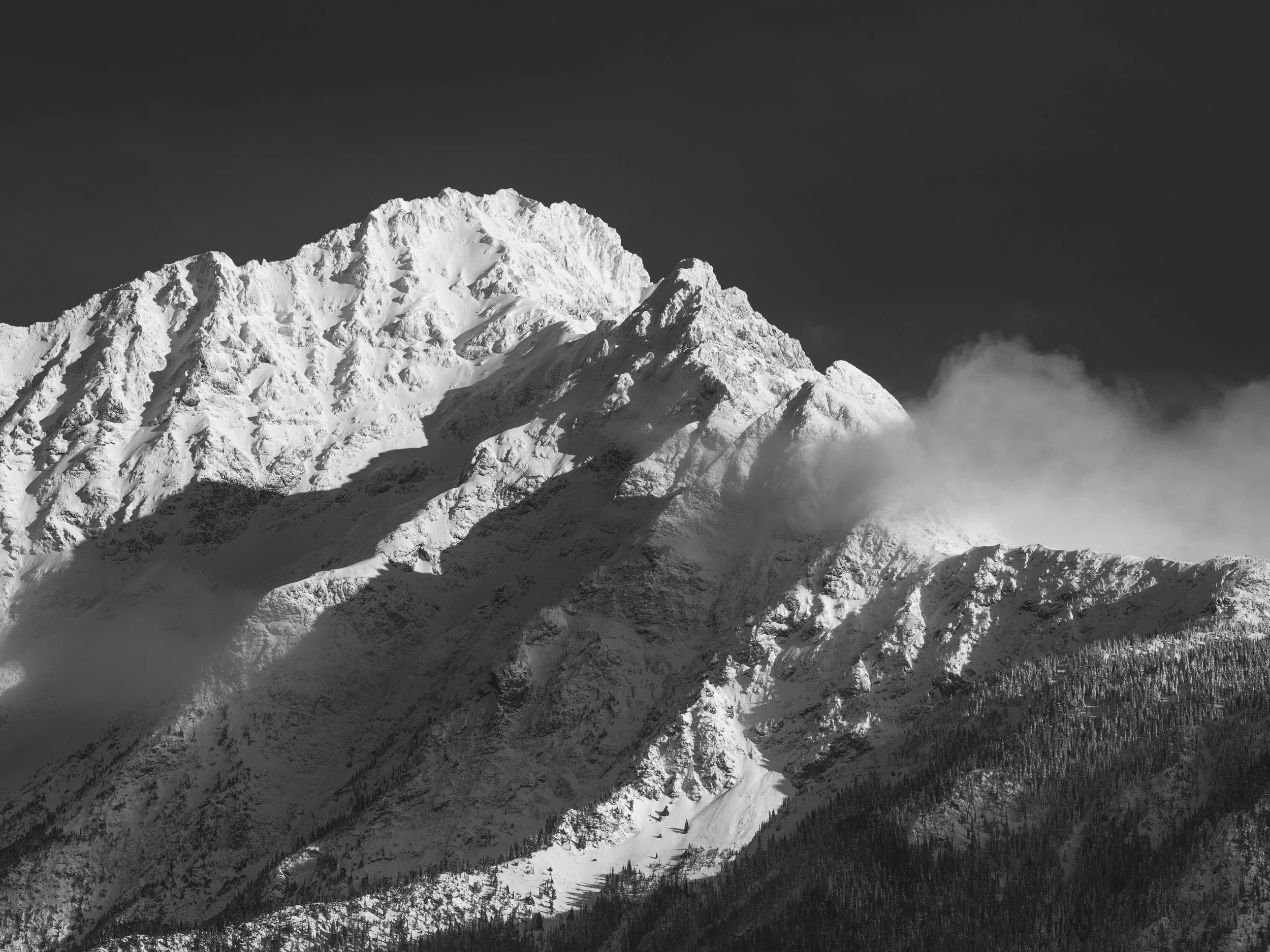 The mountain presents a complex upper ridge with multiple summits and connecting saddles, the geometry legible in fine detail because the light is direct and the air is clear. A shadow falls diagonally across the left-center of the face — the shadow 