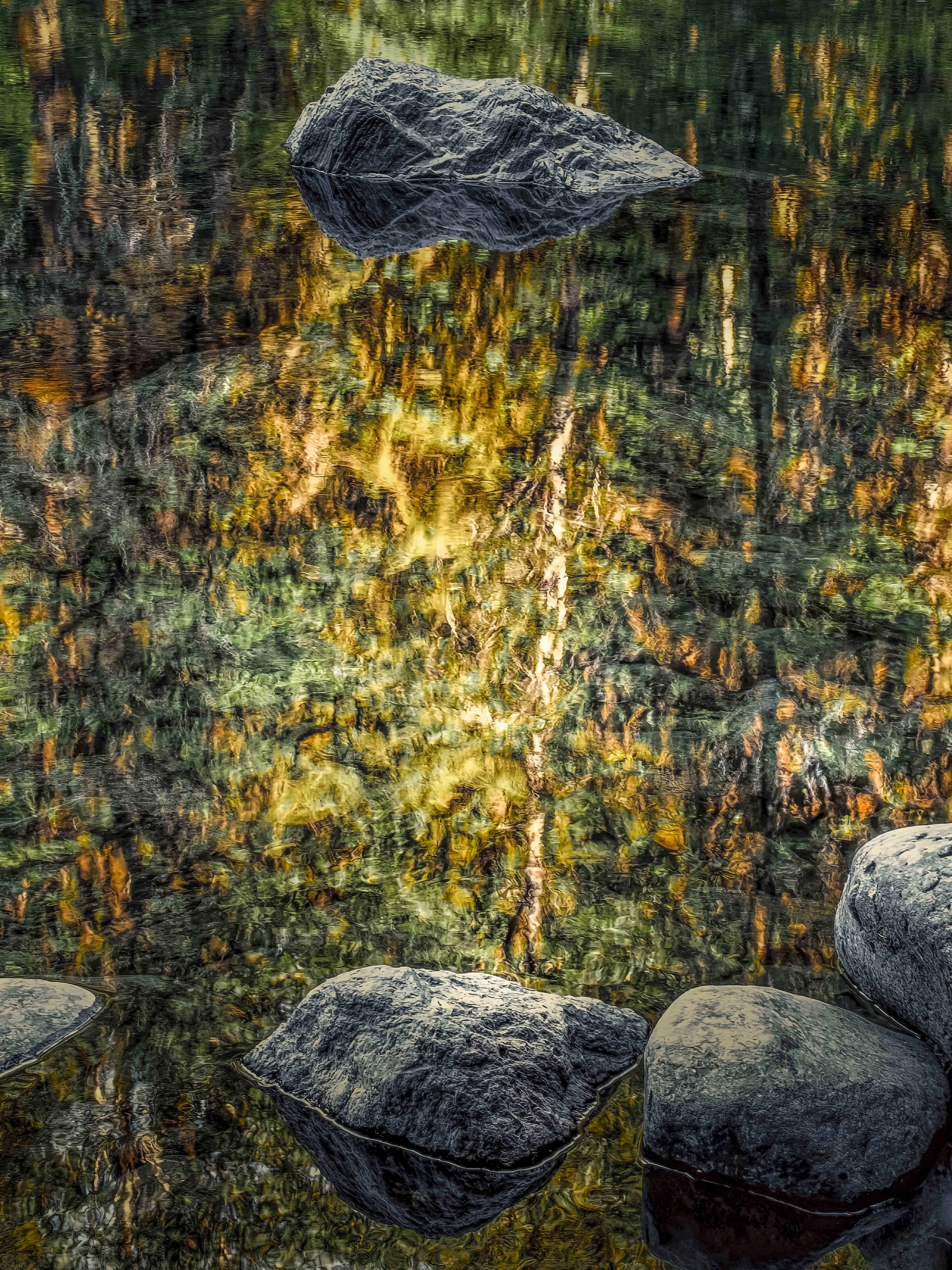 The central paradox is what arrests you immediately. The rocks appears to be floating in mid-air. The water around them is so filled with reflected autumn color — the golds, oranges, and greens of the surrounding forest — that it reads as a luminous,