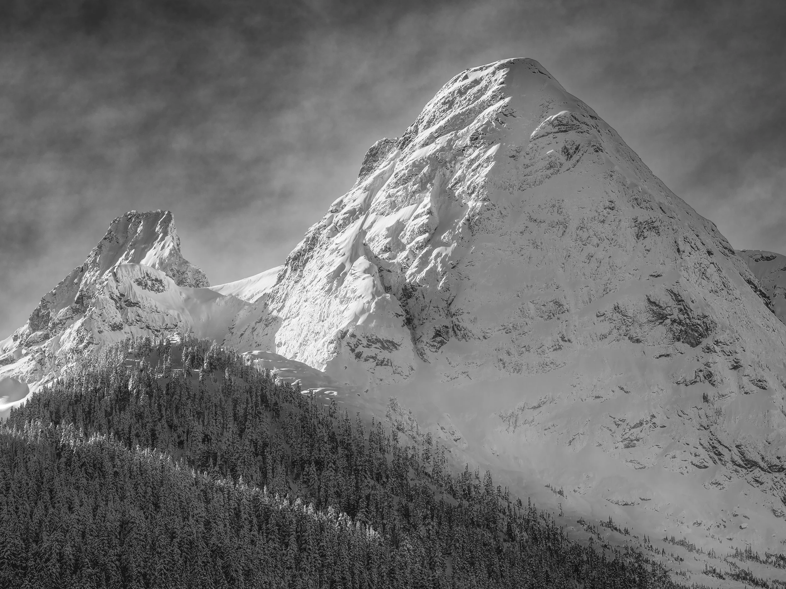 Two peaks occupy the frame in a relationship of scale and form that the eye immediately reads as a conversation: on the left, a smaller and more pointed summit — angular, jagged, its narrow ridge dropping steeply from both sides — and on the right, t