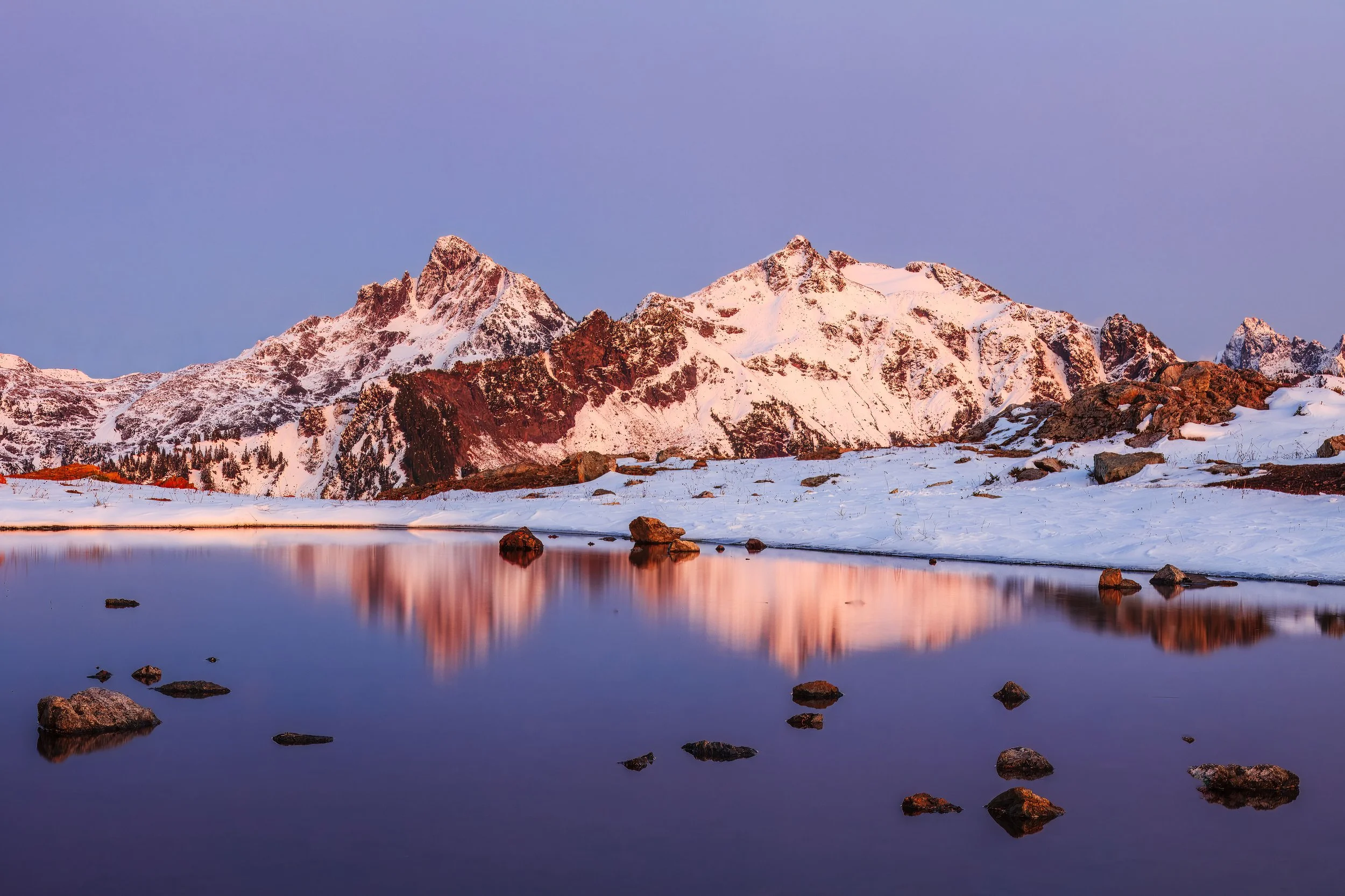 This is textbook alpenglow — that brief window just after sunset (or just before sunrise) when the sun is below the horizon but still lighting the highest peaks from below. The warm coral and salmon tones raking across the snowfields contrast beautif