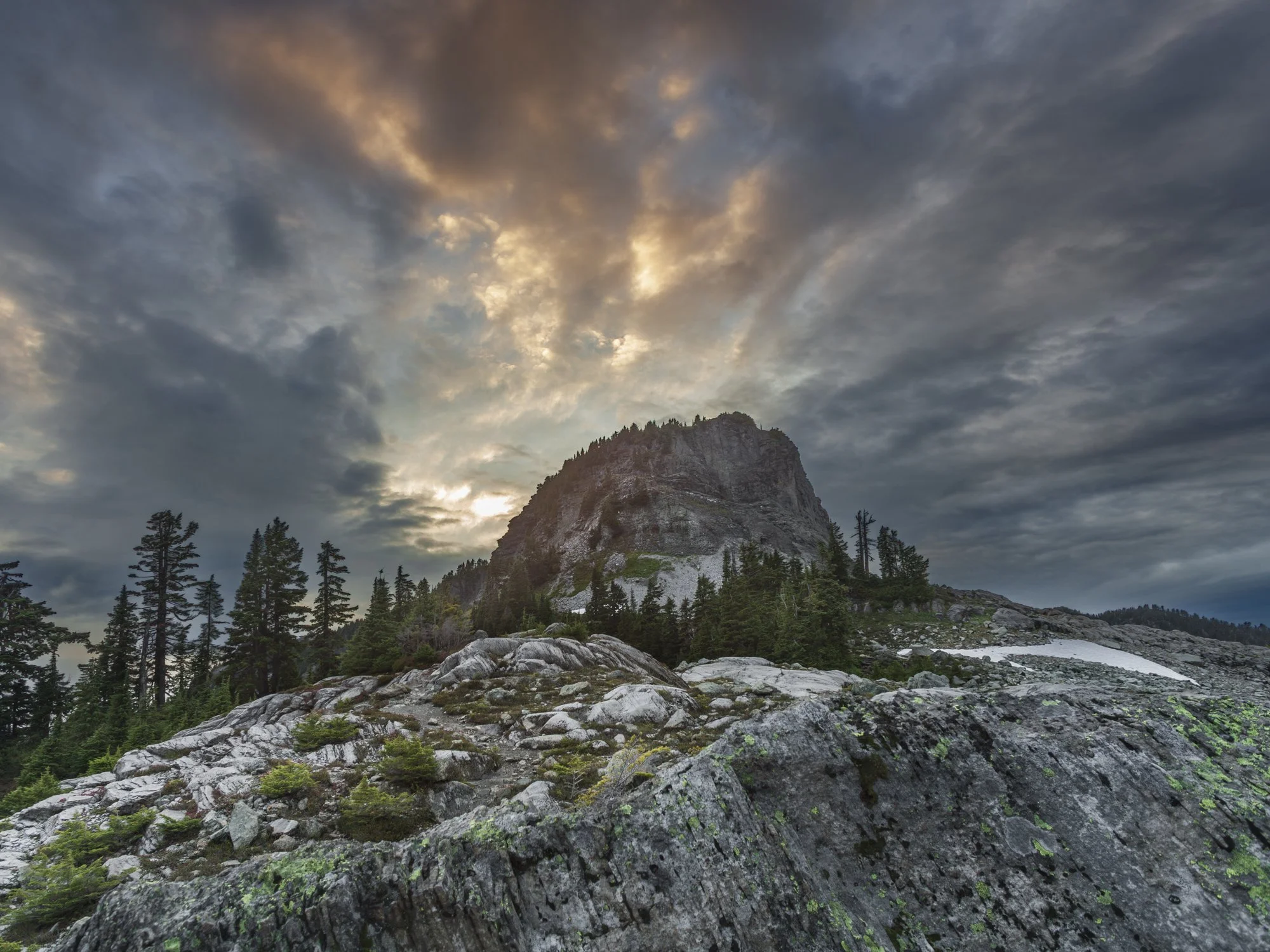 Table Mountain — that distinctive flat-topped basalt butte above Heather Meadows — caught it in genuinely dramatic conditions.

The low camera angle is the defining compositional choice. By getting down close to the lichen-covered granite slabs in th