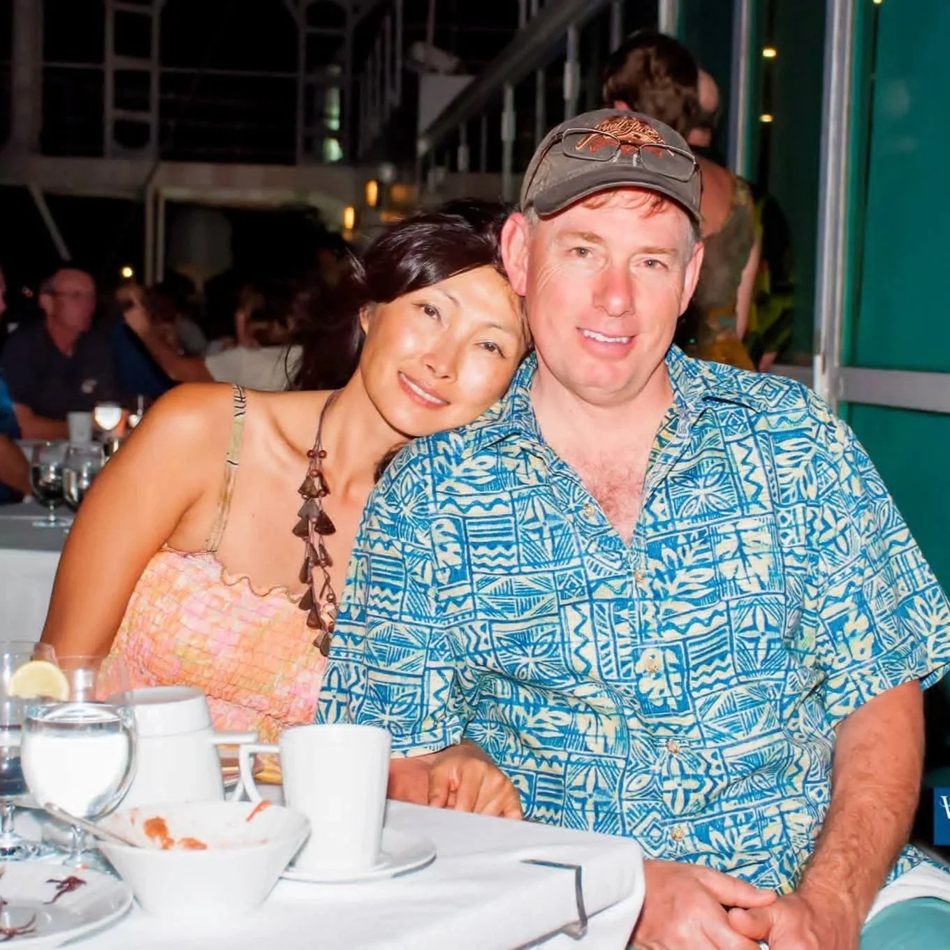 A man and a woman sitting close together at a table in a restaurant, smiling at the camera. The woman is leaning her head on the man's shoulder, wearing a sleeveless top and a necklace, while the man is wearing a blue Hawaiian shirt and a cap with glasses on it.