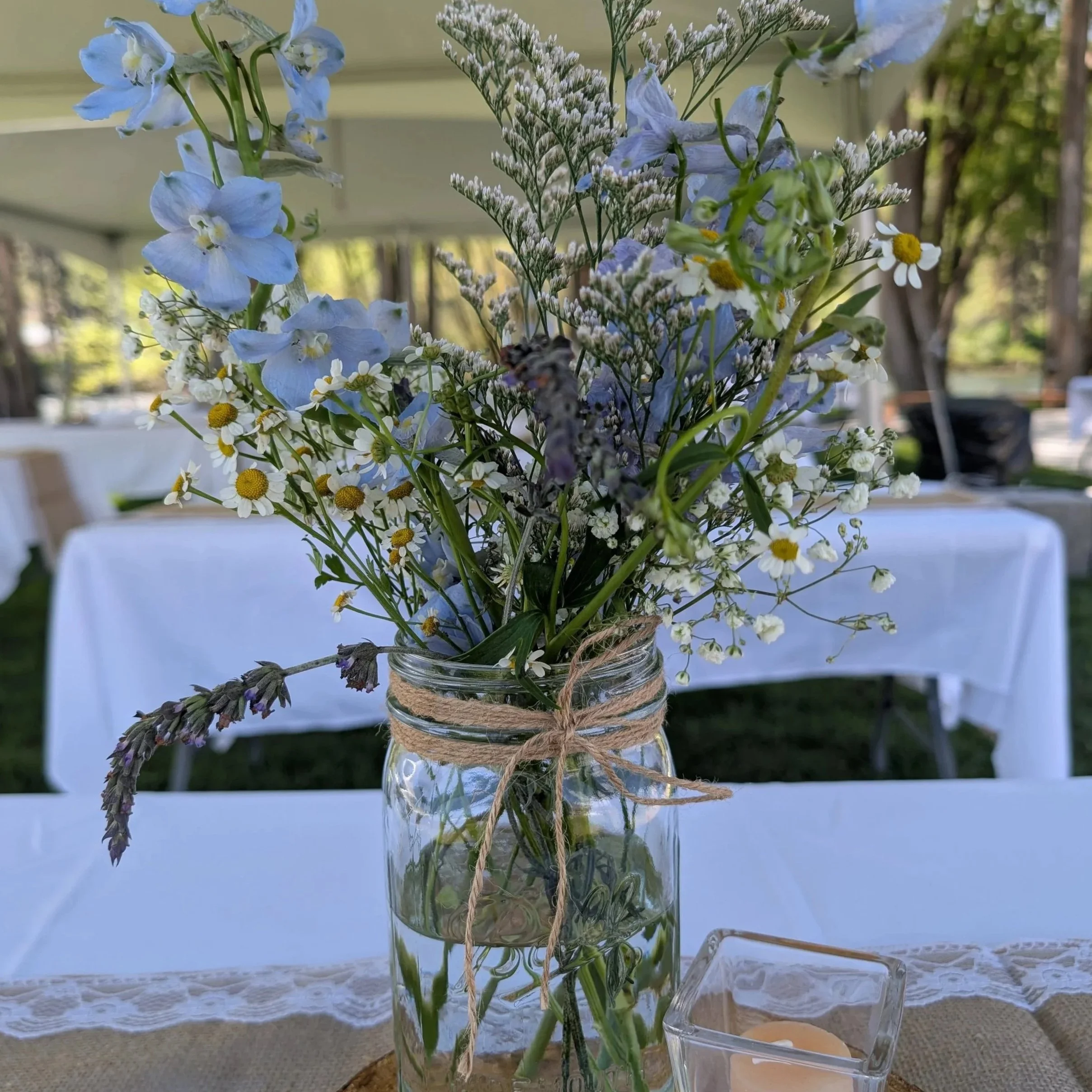 A clear glass jar with a bouquet of blue, white, and yellow flowers, including daisies and other wildflowers, wrapped with twine. A bumblebee is flying near the flowers, and a blurred outdoor setting is visible in the background.