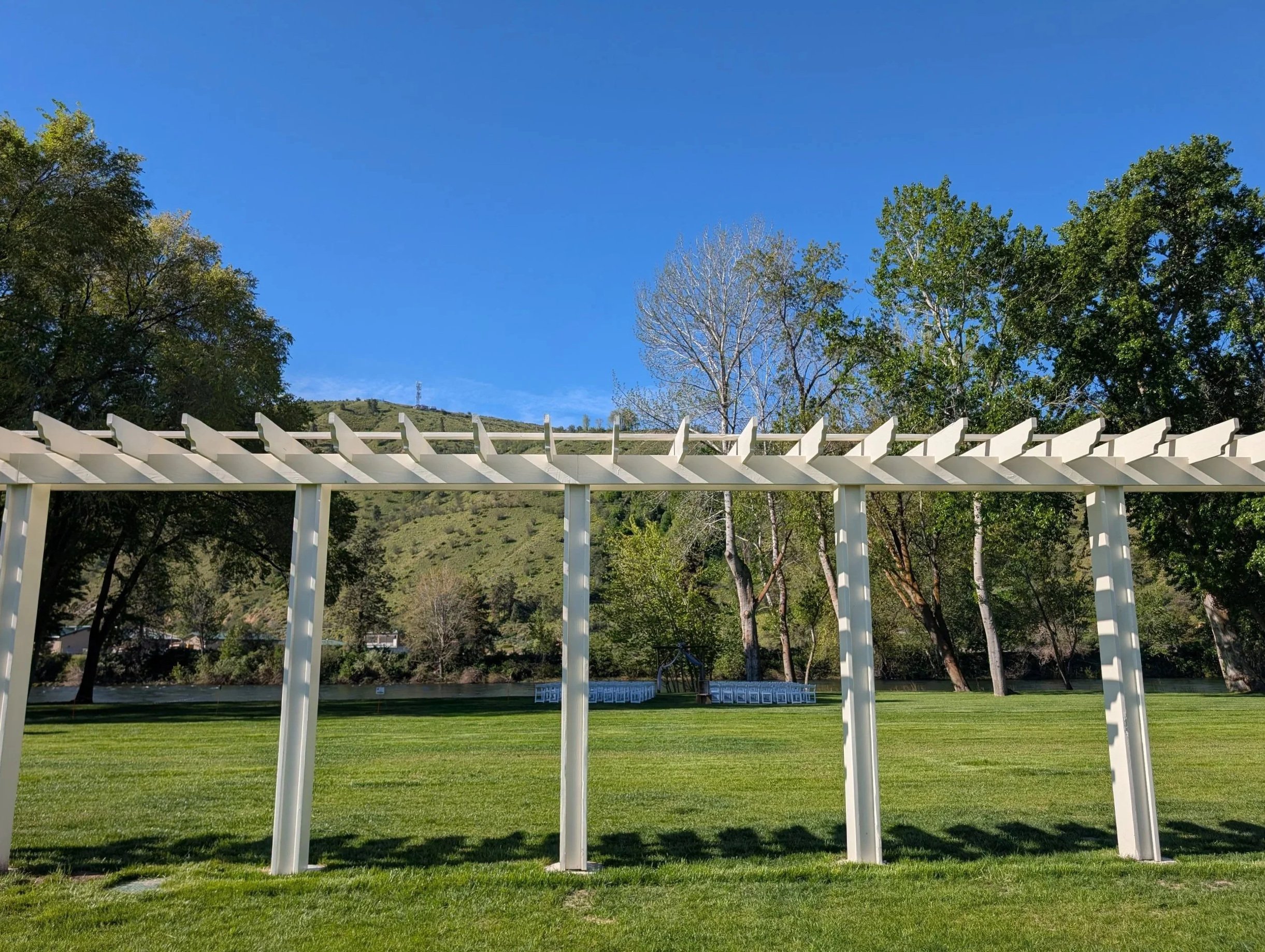 A white pergola with a grassy lawn underneath, trees in the background, and a clear blue sky above.