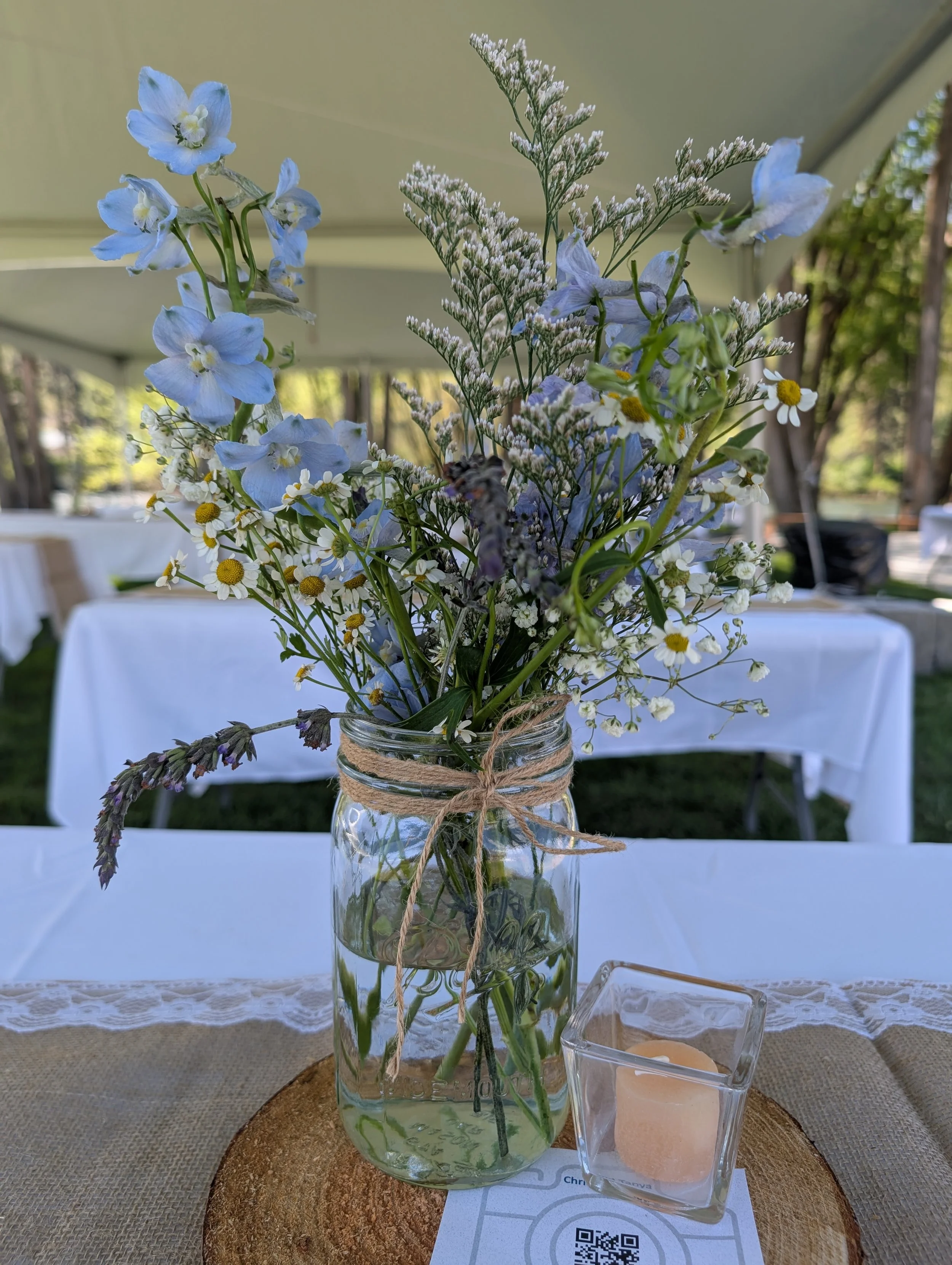 Table centerpiece of a mason jar with blue and white flowers and greenery, on a wooden plank with a candle in a glass holder and a card with a QR code.