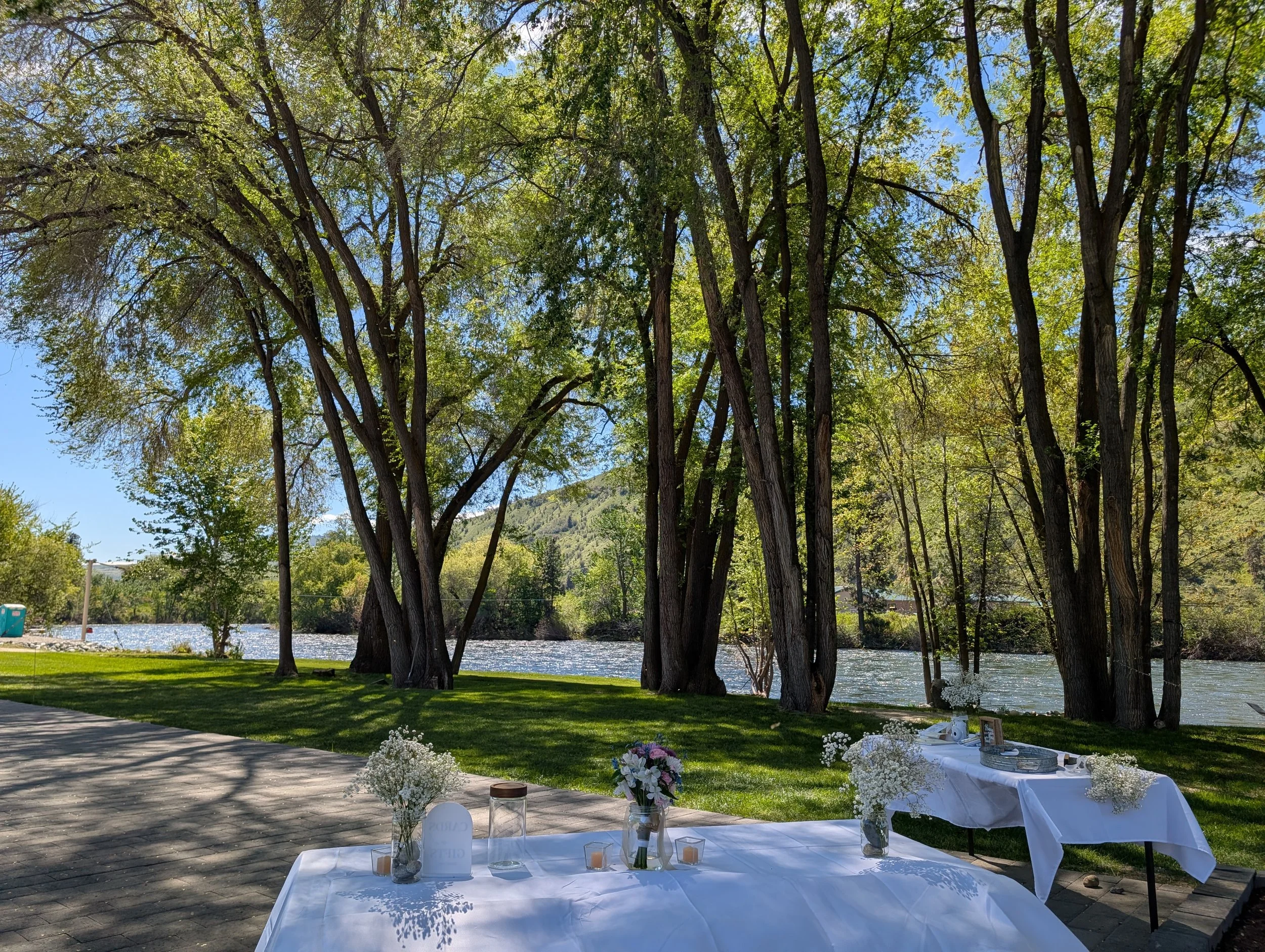 Outdoor wedding reception setup near a river, with a long white table decorated with flowers and candles, under tall green trees on a sunny day.