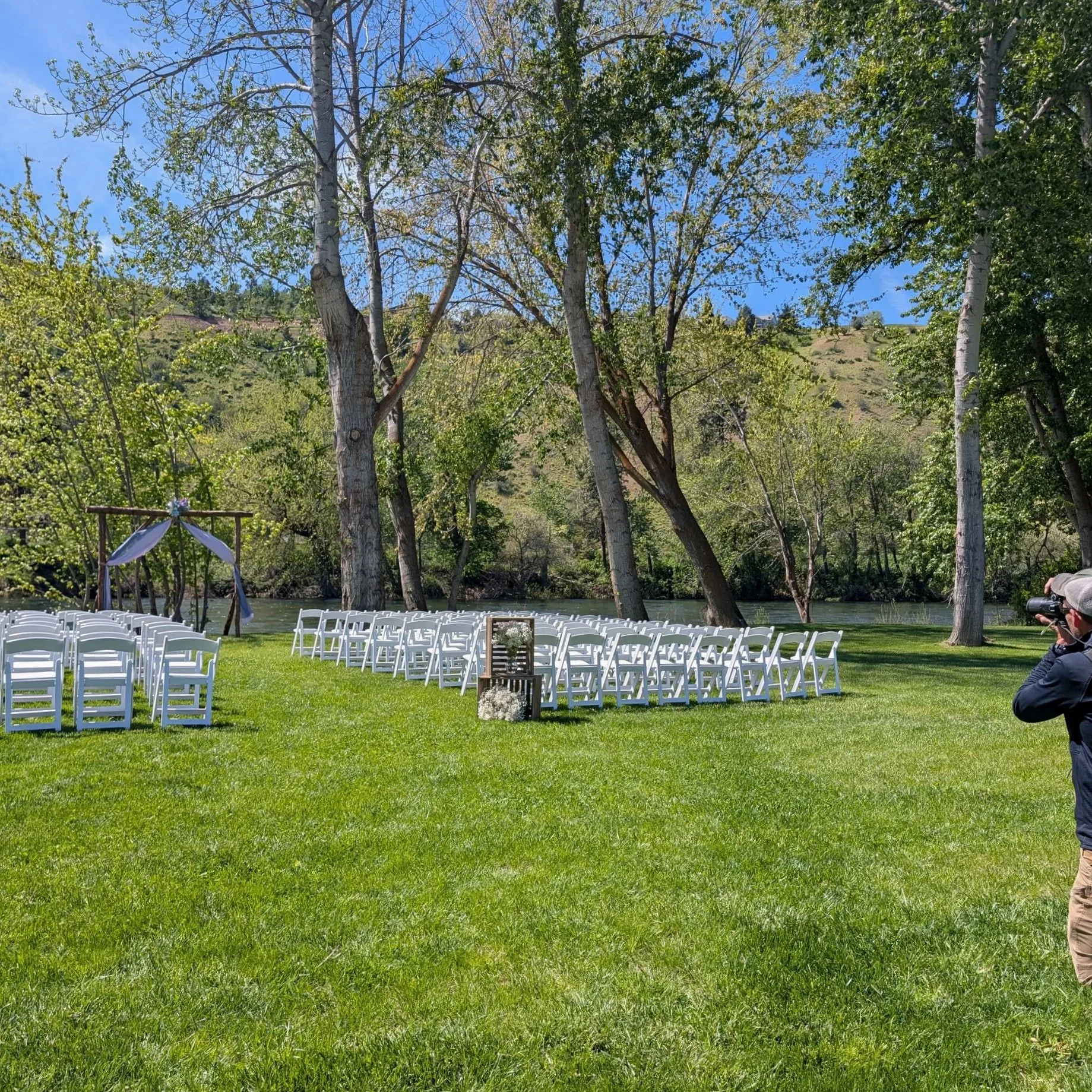 Outdoor wedding setup with white chairs arranged in rows on a lush green lawn, overlooking a river and surrounded by trees, with a hillside in the background under a partly cloudy sky.