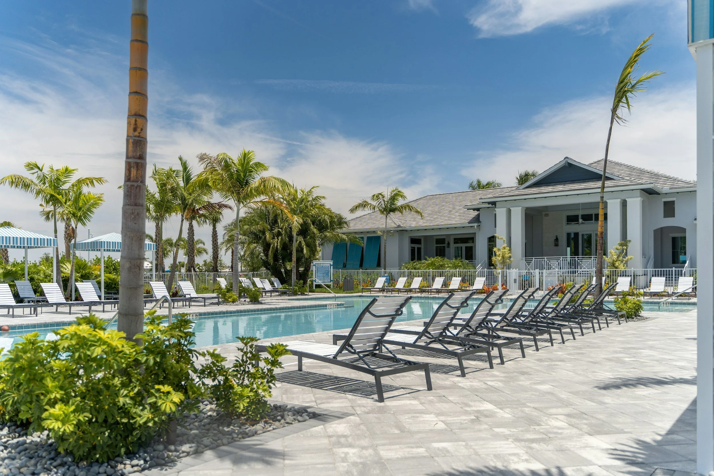 Swimming pool area with lounge chairs, umbrellas, and palm trees near a modern building under a partly cloudy sky.