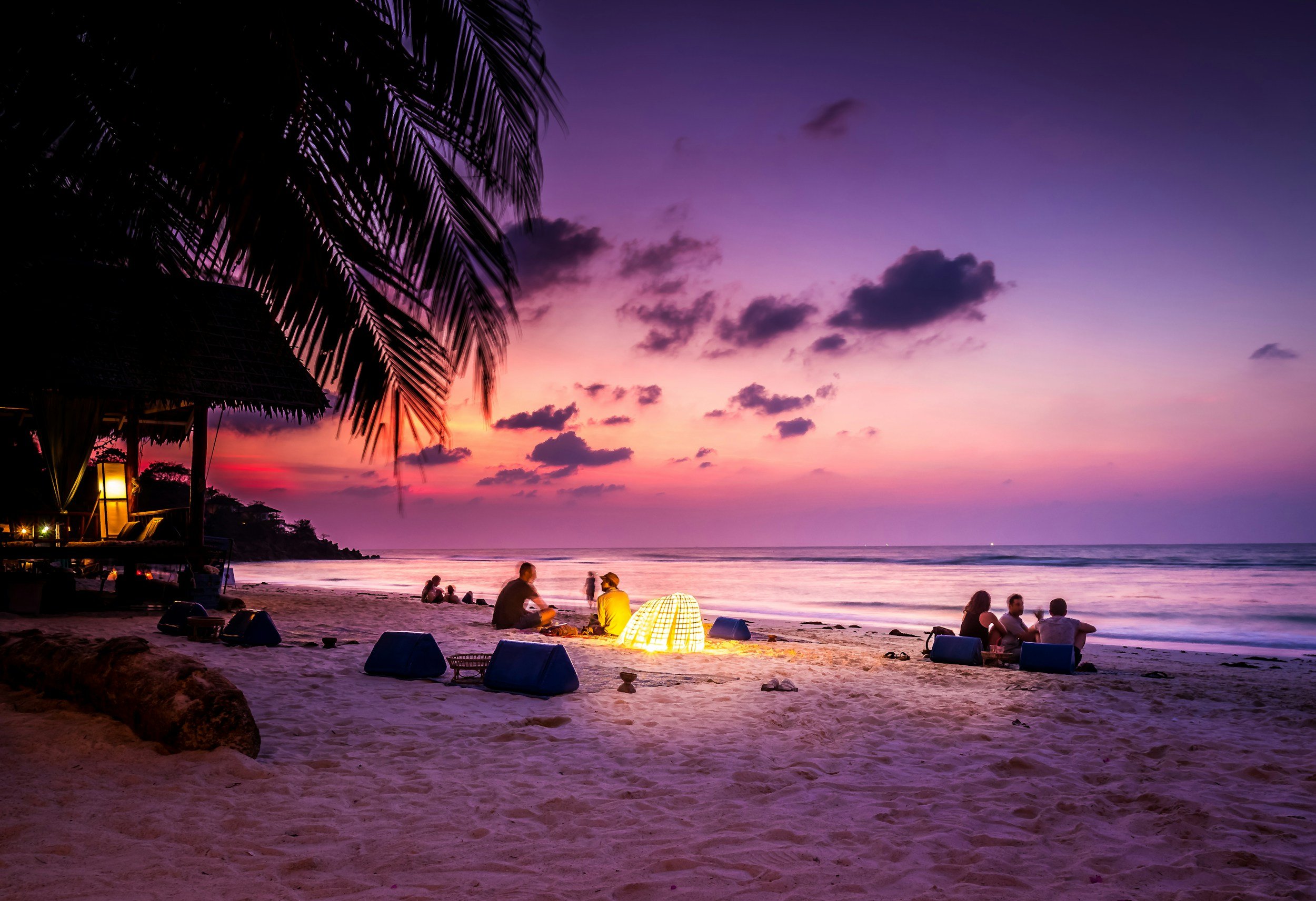 People sitting on the beach during sunset, with purple and pink sky, palm trees, and a thatched hut with a lantern