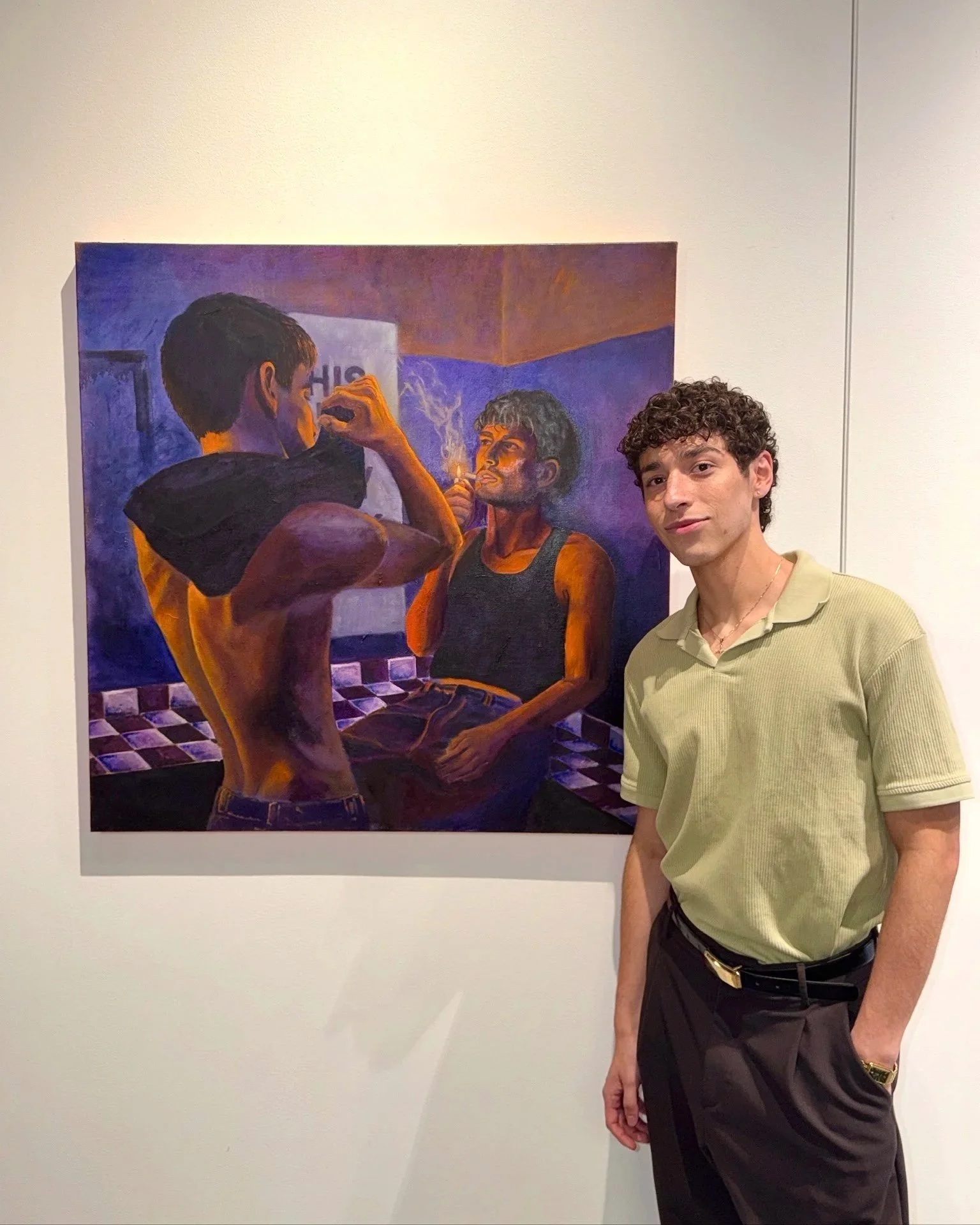 A young man with curly hair, wearing a light green polo shirt and dark pants, stands beside a painting of two men, one standing and one sitting, in a gallery with white walls.