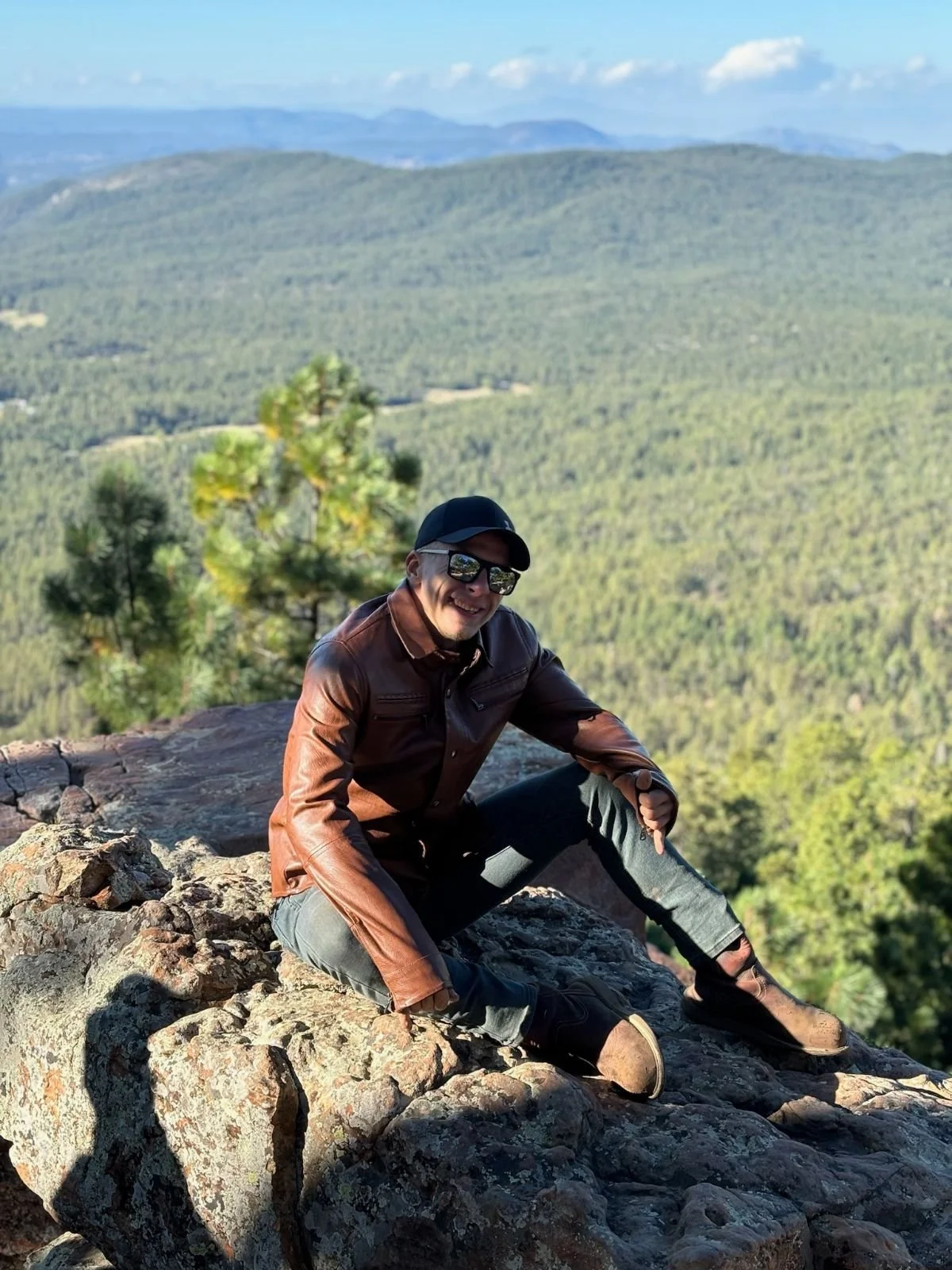 PiPPuP's Founder, A Caucasian man in a leather jacket, black hat, and sunglasses, sitting on cliffside edge rocks with a scenic forested mountain valley in the background.
