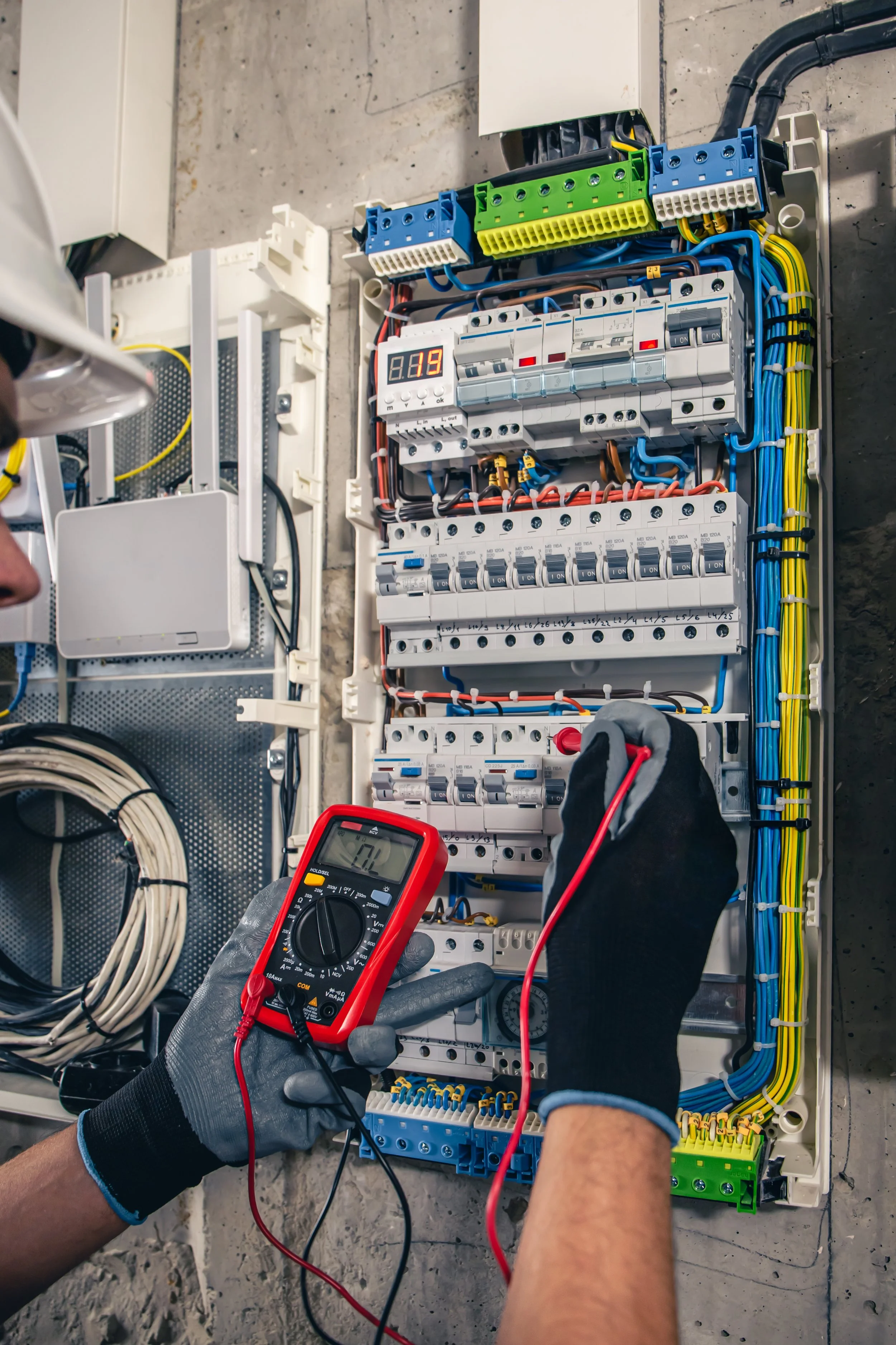 Person testing electrical panel with multimeter. Open electrical panel with wires, circuit breakers, digital display, and various electrical components. Worker wearing gloves and holding multimeter probes.