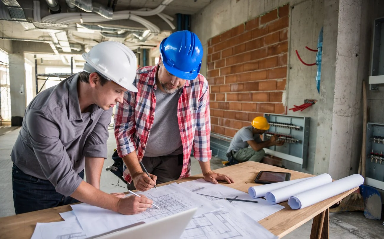 Two male engineers wearing hard hats working on blueprints at a construction site with a third worker in the background managing electrical wiring.