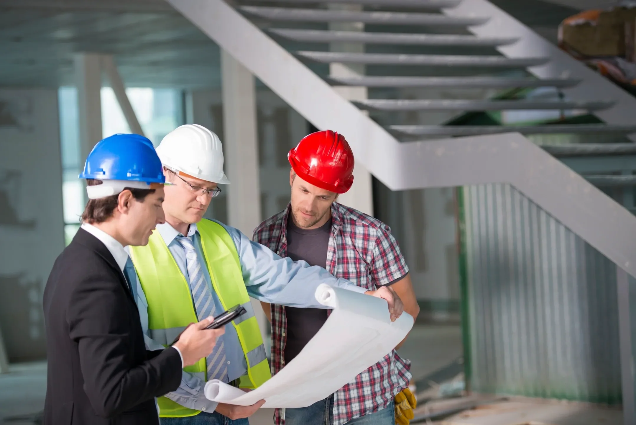 Three construction workers wearing hard hats and safety vests examining blueprints inside a building under construction.