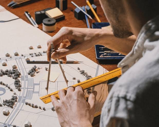 Architect or urban planner working on a detailed scale model or map with a compass and a ruler, surrounded by drafting tools and materials on a wooden desk.