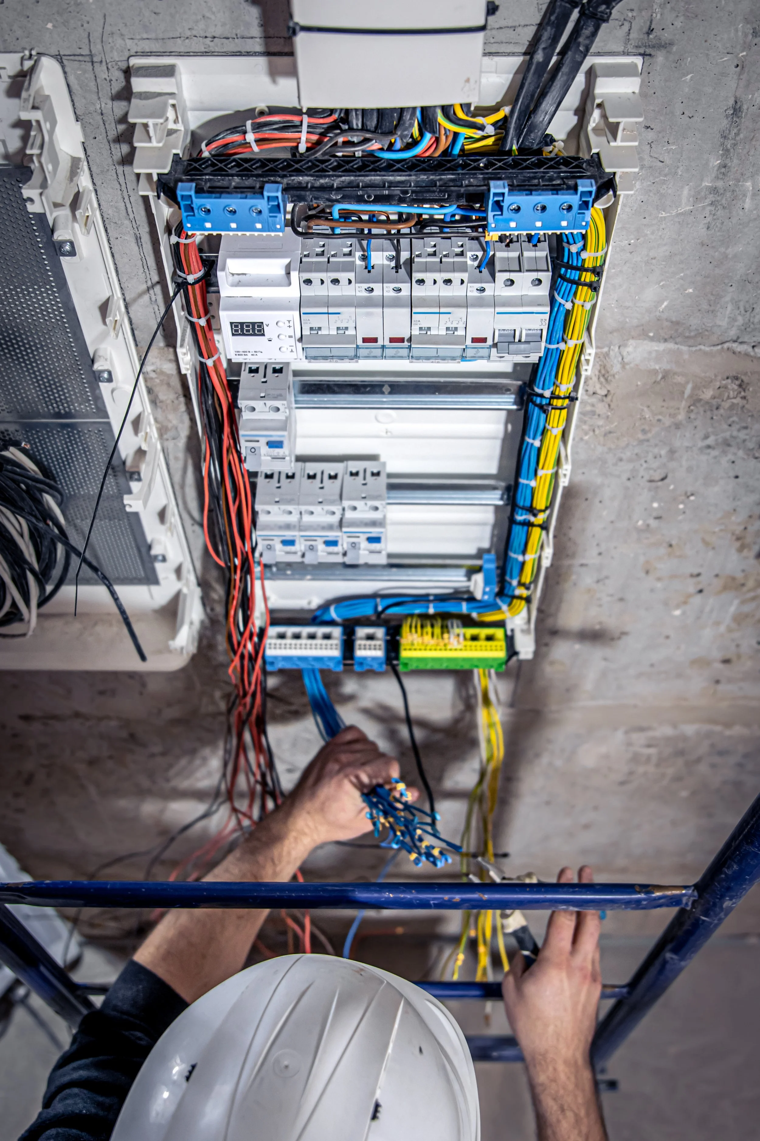 Technician working inside an electrical control panel, connecting blue wires with a screwdriver, with a white part of safety helmet visible at the bottom of the image.