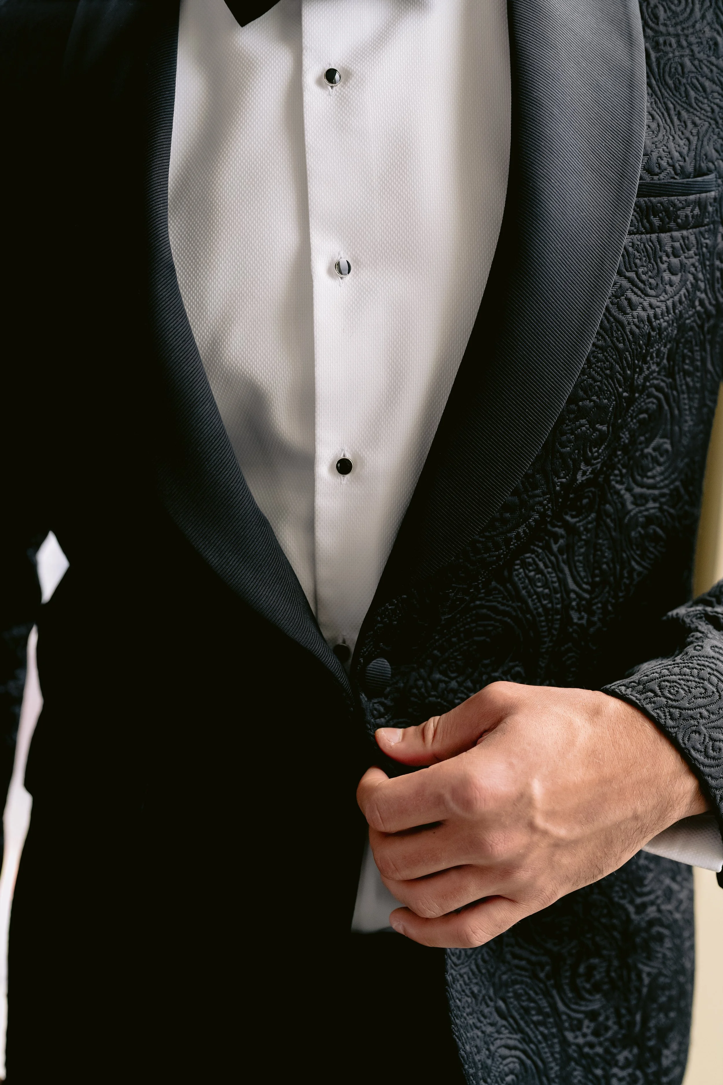 Close-up of a man in a tuxedo with a textured dark jacket and white shirt, holding his hands crossed in front of him.