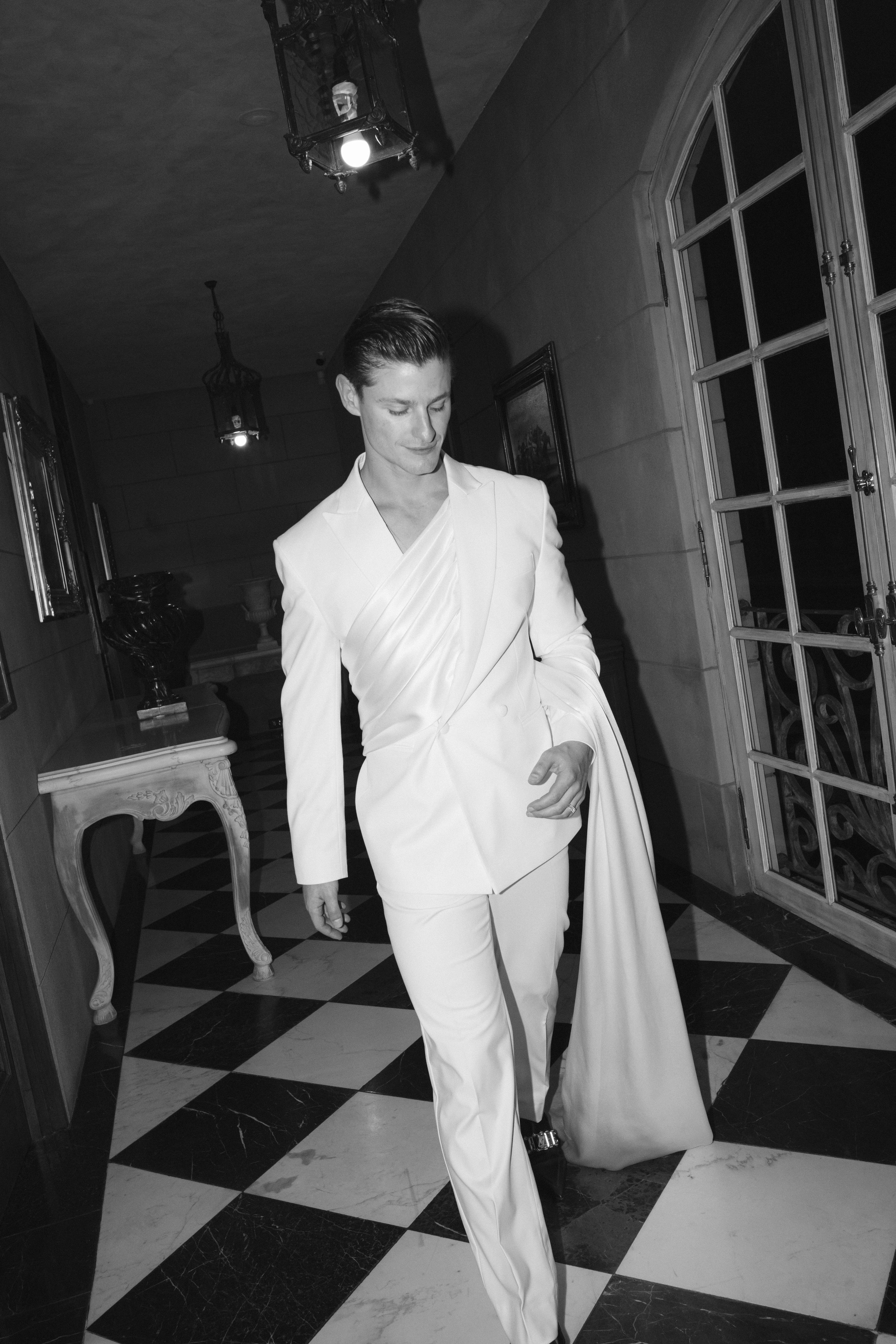 A man wearing a white suit walking in a decorated hallway with checkered floor, ornate furniture, framed artwork, and hanging lantern-style lights.