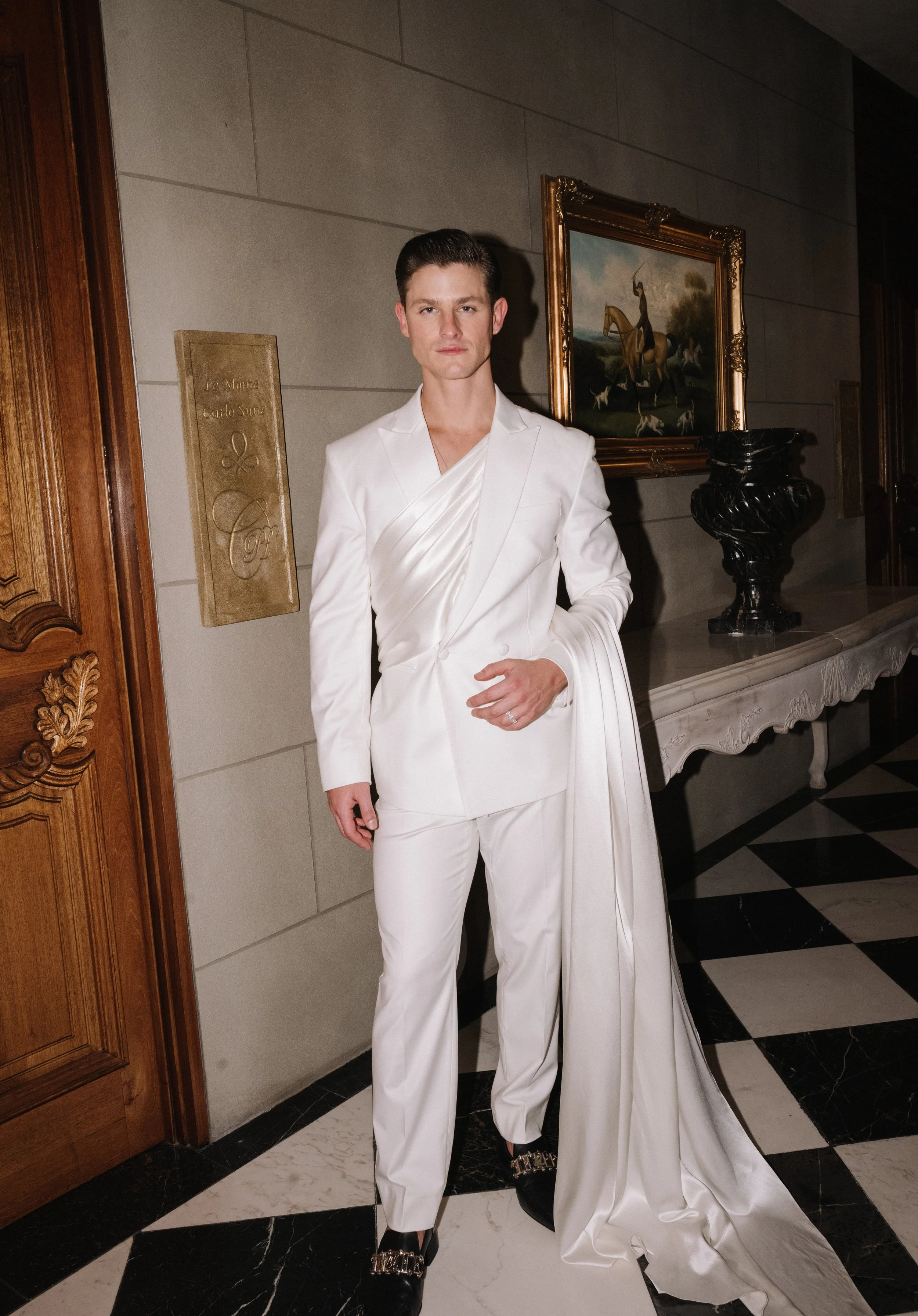 A man dressed in a stylish white tuxedo with a satin lapel and matching pants, standing in an elegant lobby with artwork and black-and-white checkered flooring.