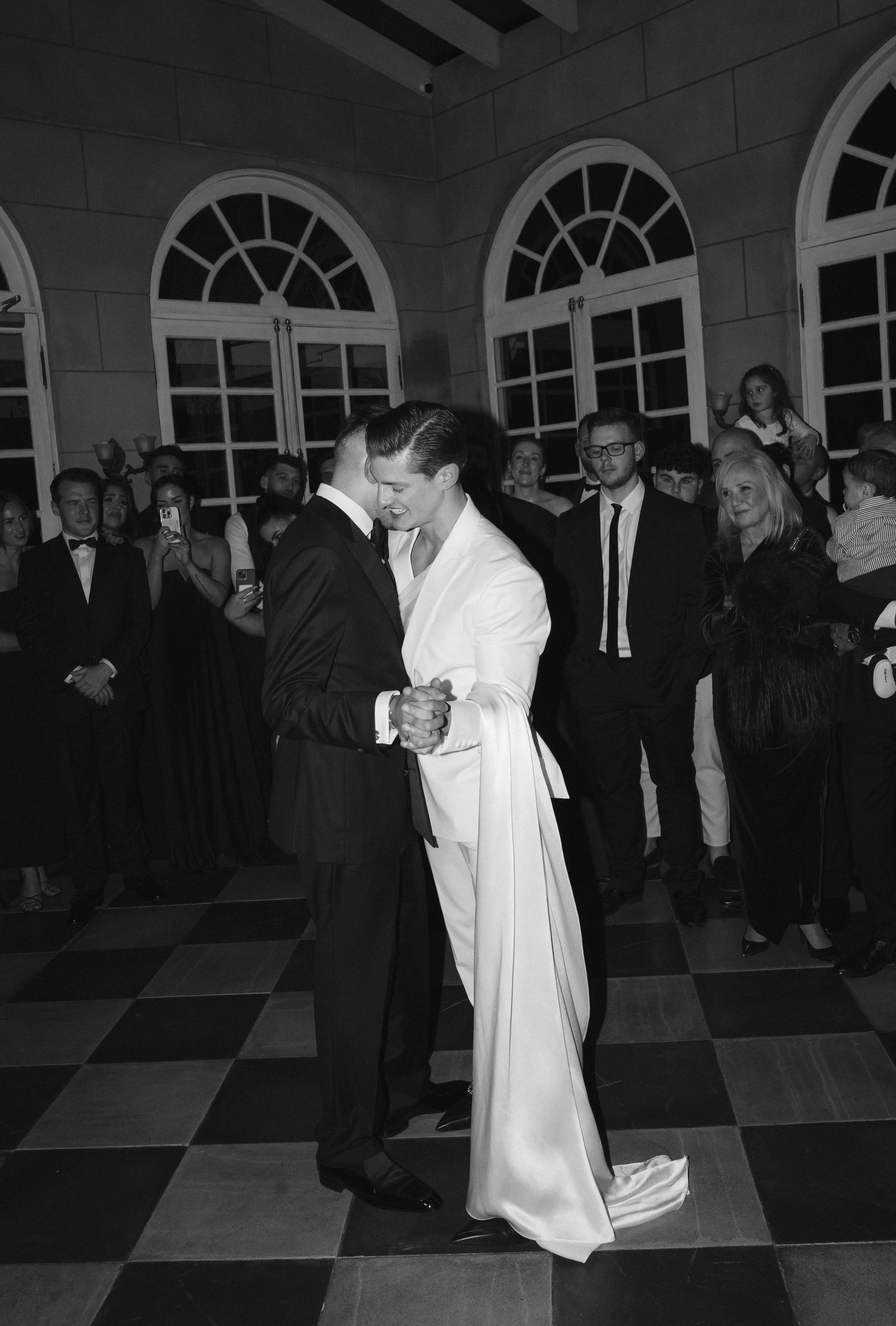 A black and white photo showing a couple dancing closely at a formal event or wedding, surrounded by guests in formal attire, inside a room with large arched windows and checkered tile flooring.
