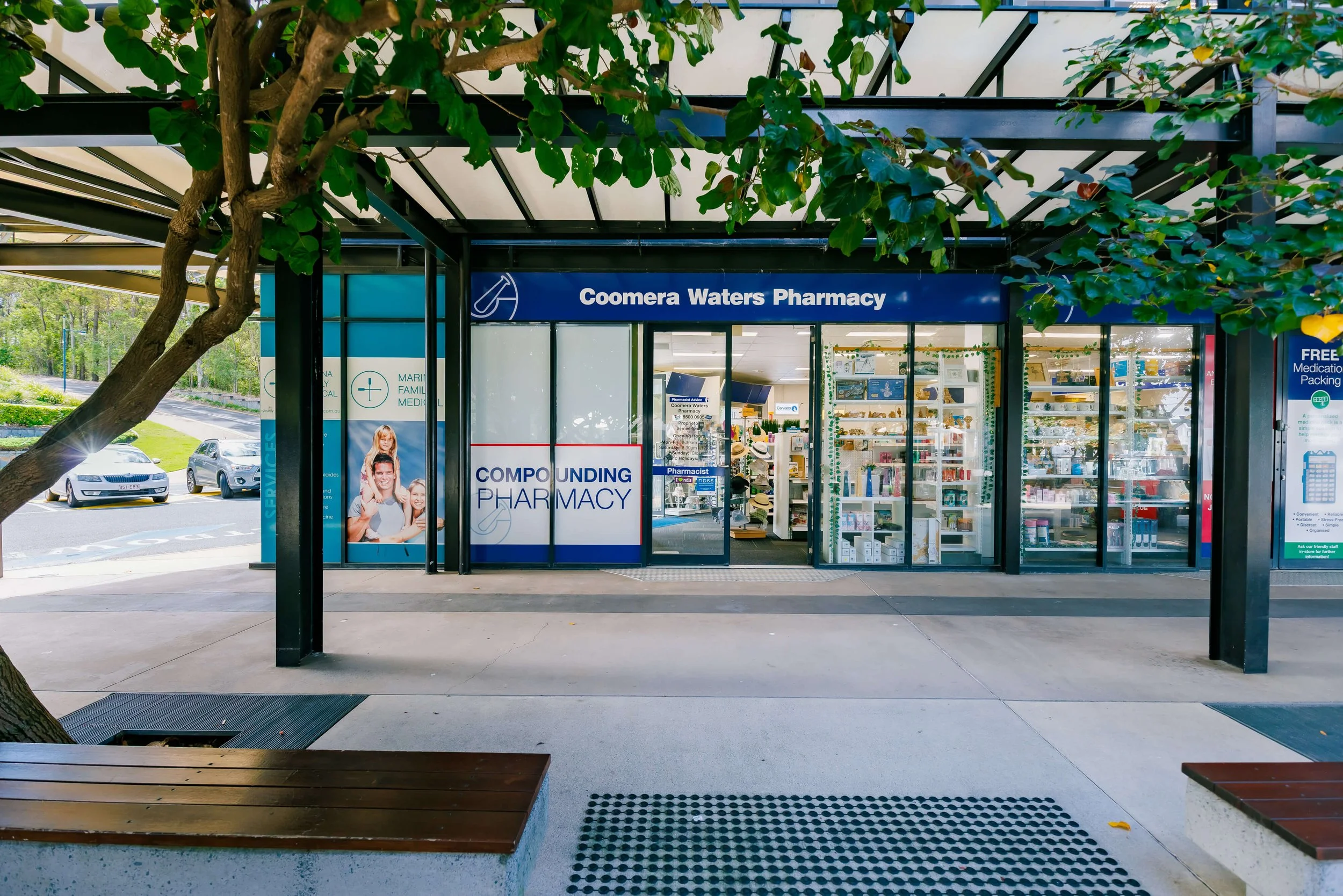 Exterior view of Coomera Waters Pharmacy with glass doors and windows displaying pharmacy products inside. A parking lot with cars and green trees in the background. 
