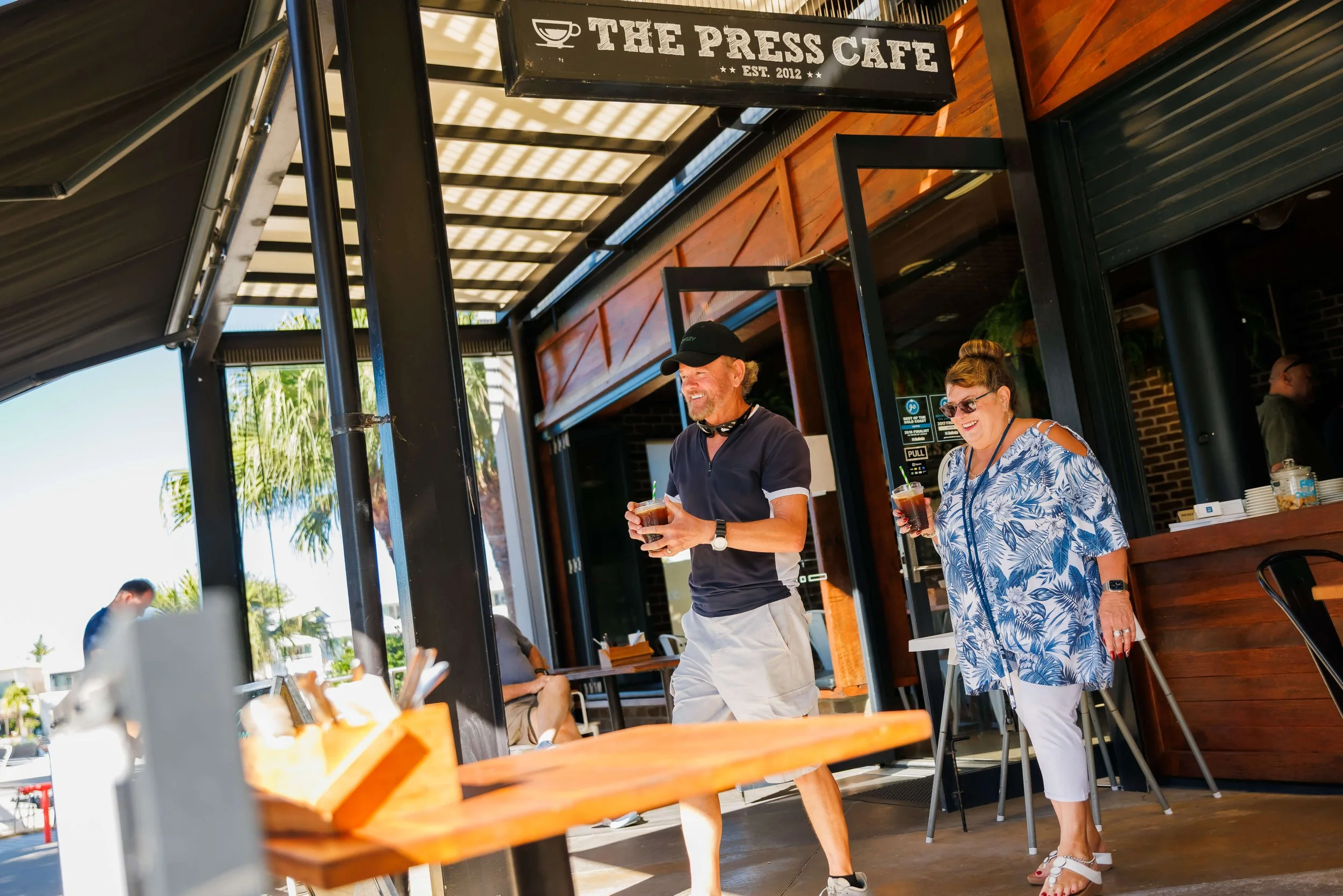 People enjoying drinks outside a cafe with a sign reading 'The Press Cafe', under a shaded awning, with palm trees in the background.